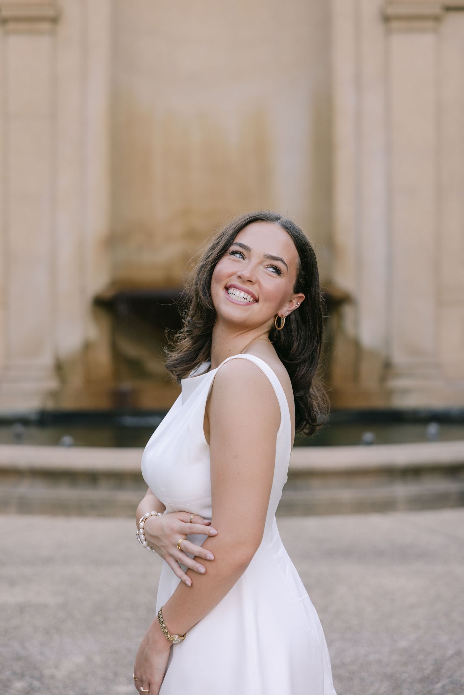 A young woman smiles over her shoulder in front of a stone fountain