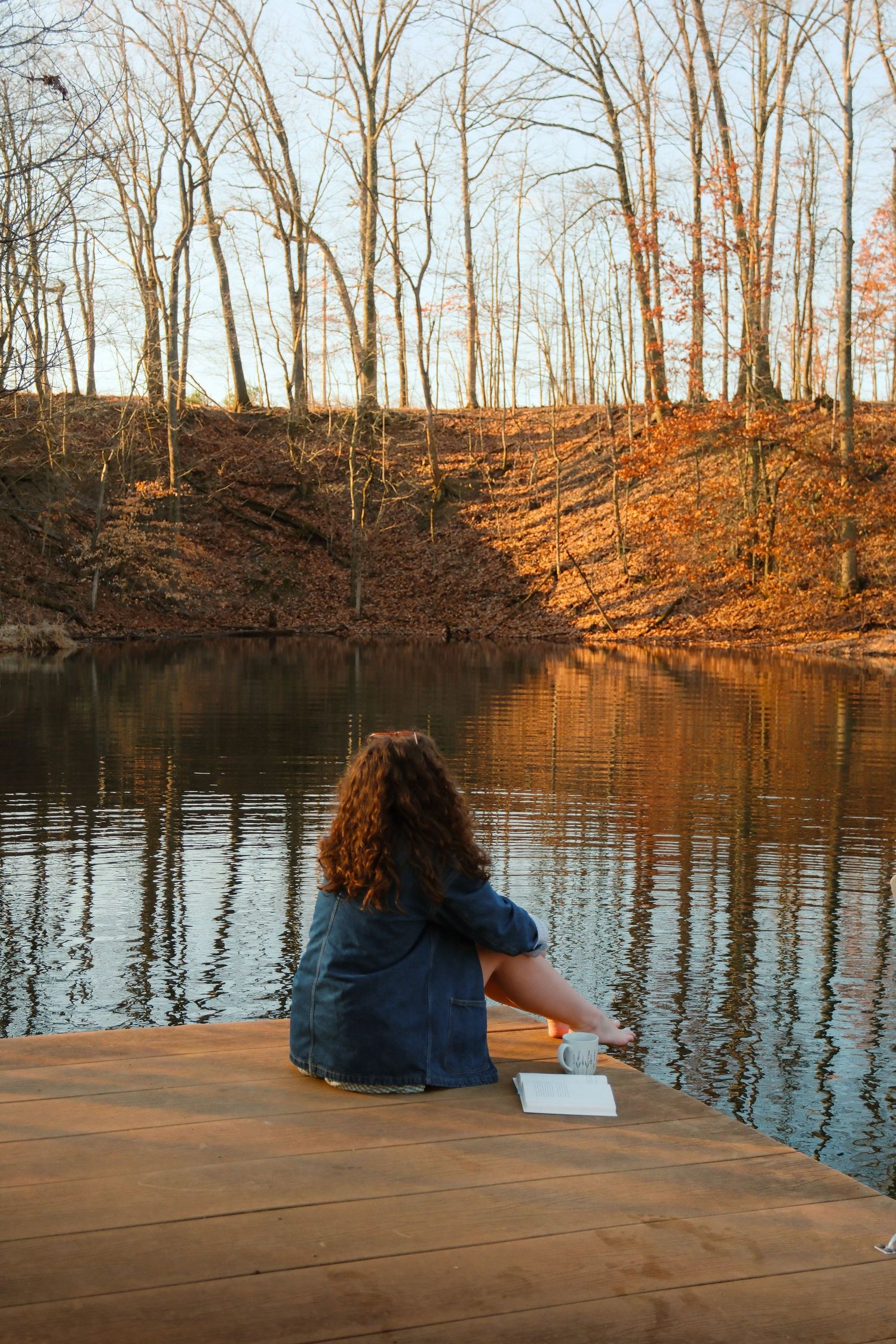 A person sits peacefully on a wooden dock by a tranquil lake surrounded by bare winter trees at sunset.