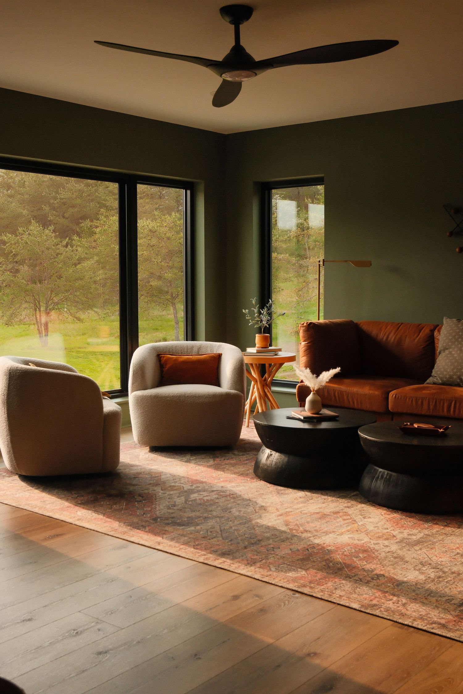Modern living room with olive green walls, rust sofa, beige armchair, black coffee table, wood floors, and large windows.