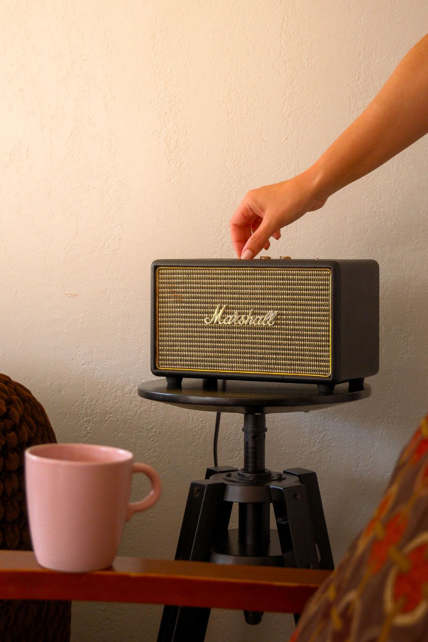 Hand adjusting knob on Marshall Bluetooth speaker on stand, with pink mug on wooden table nearby.