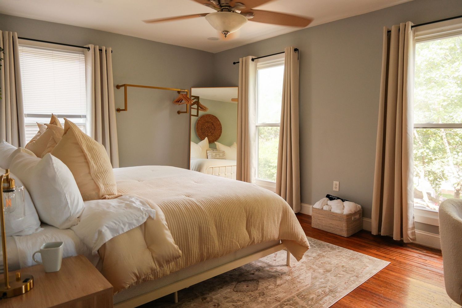 Cozy bedroom with beige bedding, hardwood floors, ceiling fan, neutral curtains, and natural light from multiple windows.