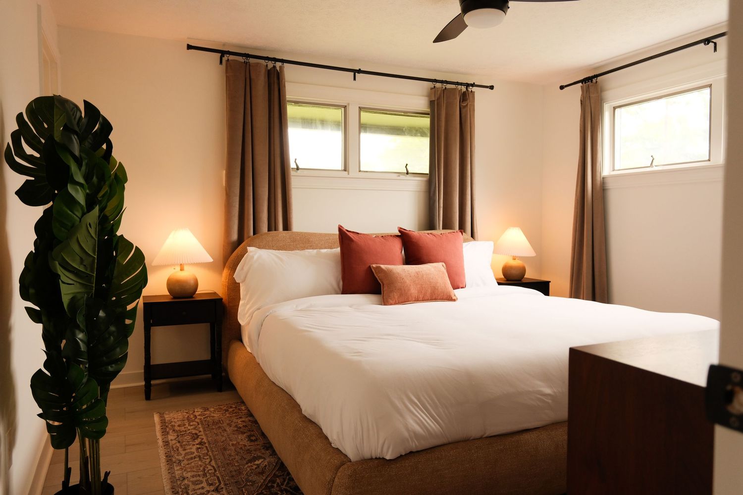 Warm bedroom with white bedding, coral pillows, tan curtains, matching nightstand lamps, and tropical plant decor.