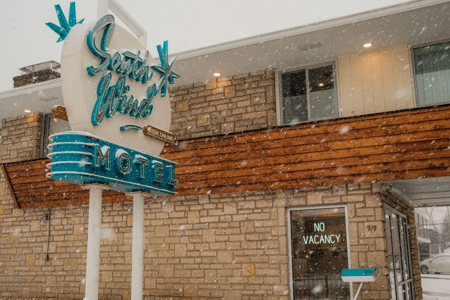 Vintage Sugar Lime Motel sign with teal lettering in falling snow, No Vacancy sign visible on brick building.