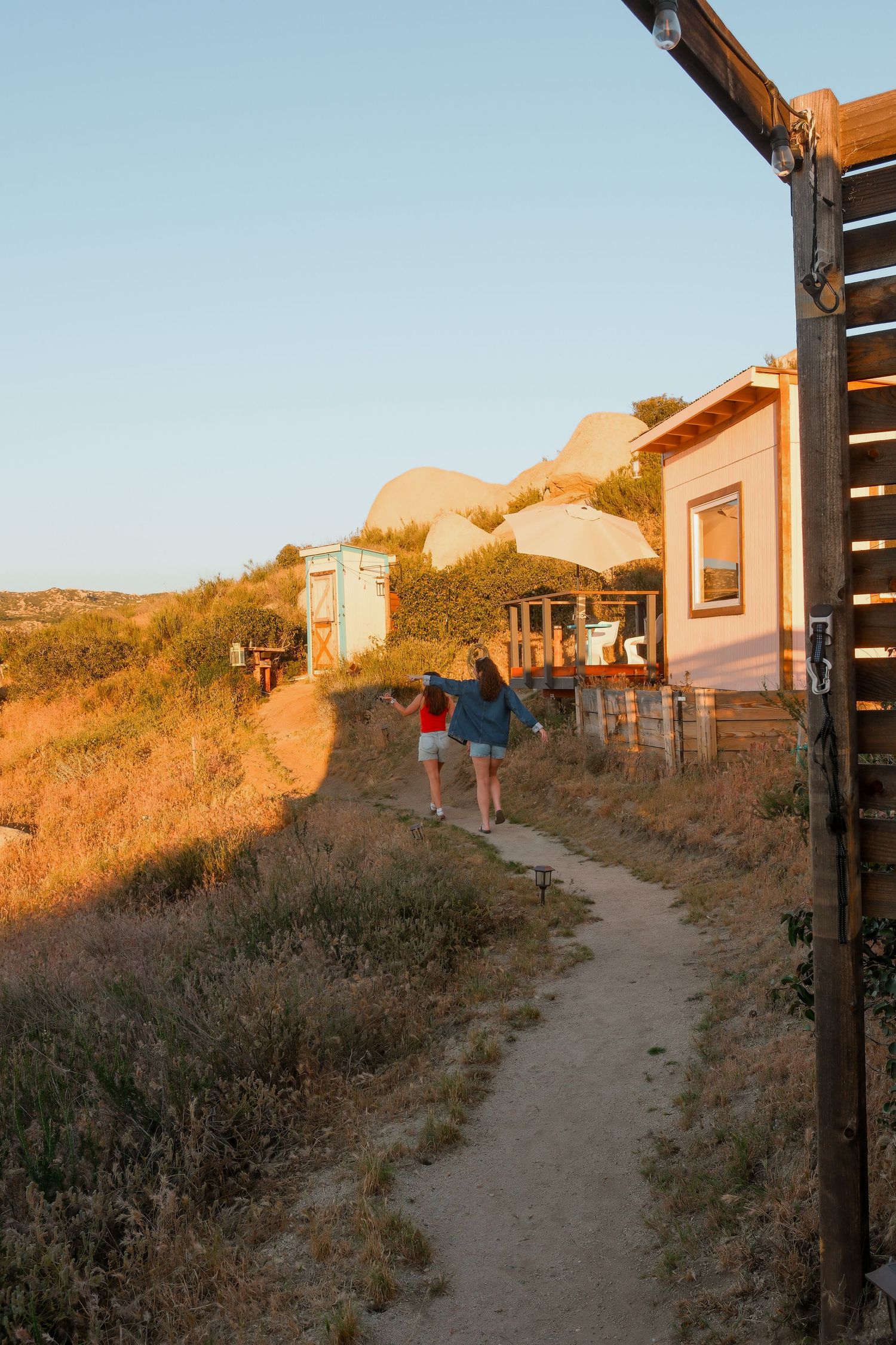 Two women walking along a dirt path past rustic cabins on a golden hillside at sunset in a remote retreat.