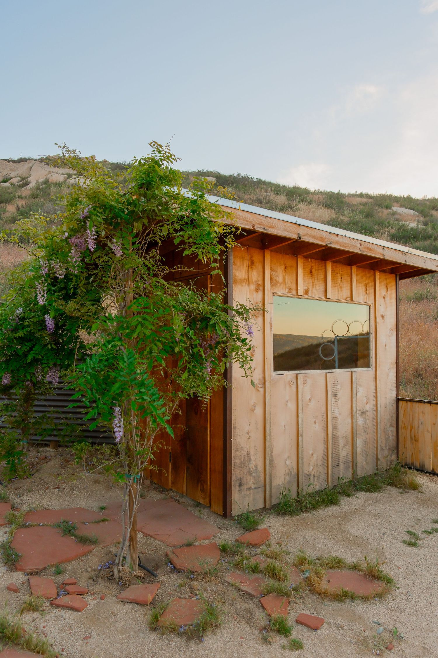 Rustic wooden cabin with a flowering wisteria tree, red stone path, and mountain backdrop at golden hour.