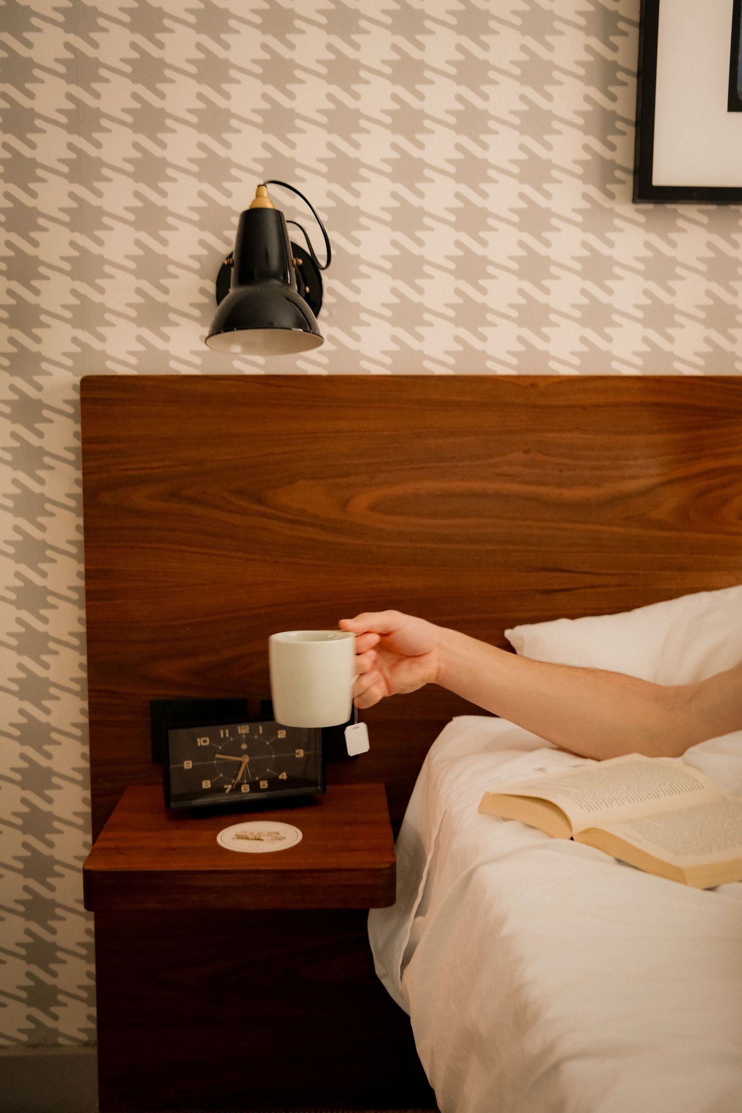 A hand reaches for a white coffee mug on a wooden nightstand beside an alarm clock in a cozy bedroom.