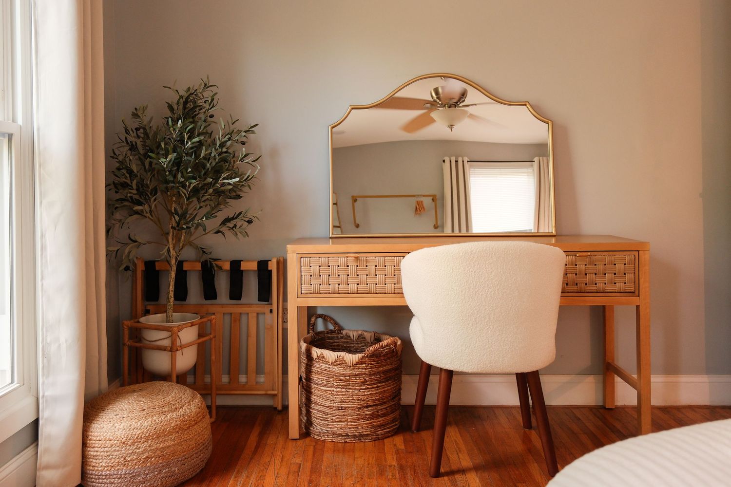 Boho bedroom vanity with rattan desk, white chair, gold mirror, woven basket, and olive tree on hardwood floors.