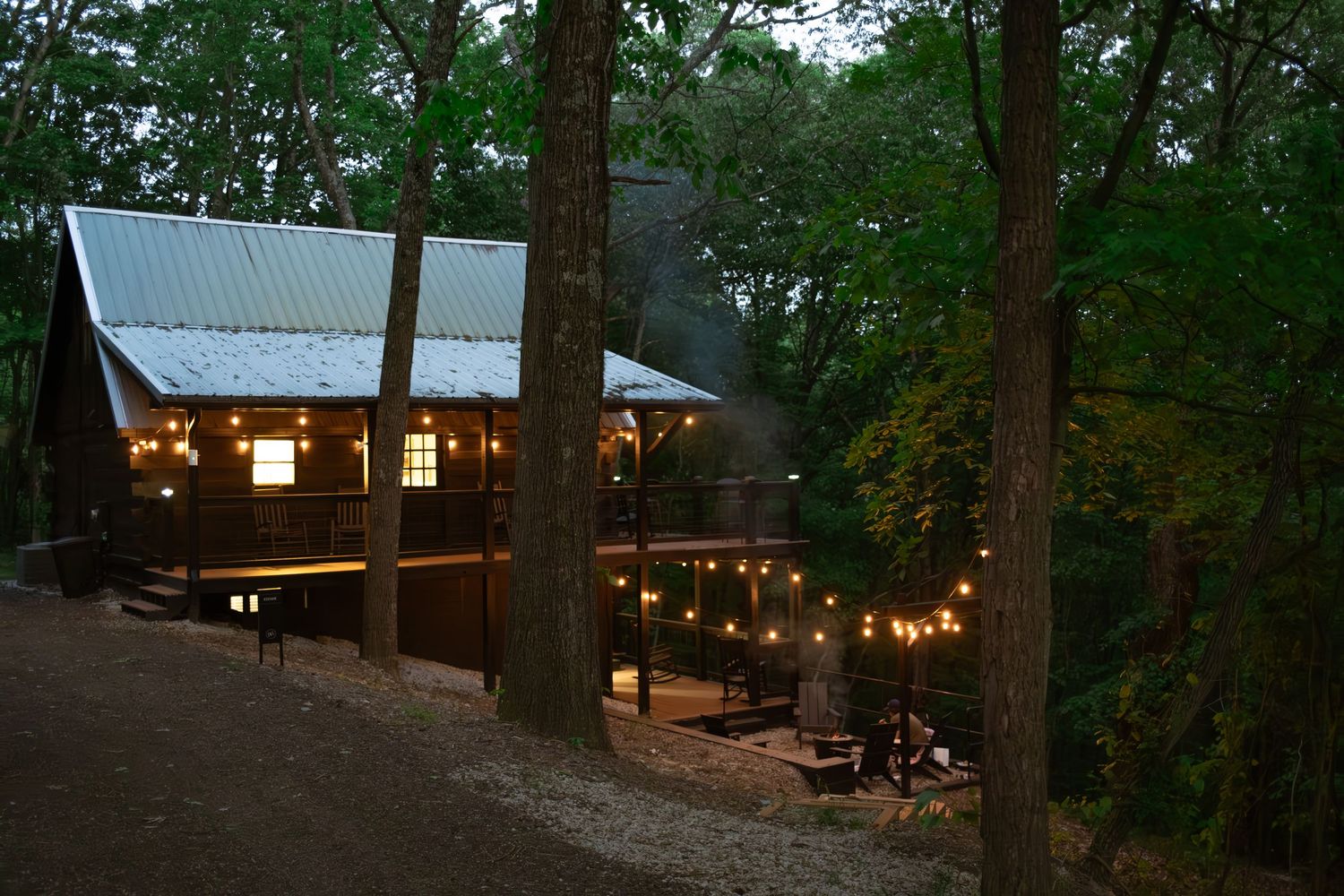 Cozy cabin deck illuminated by string lights at dusk in a wooded setting.