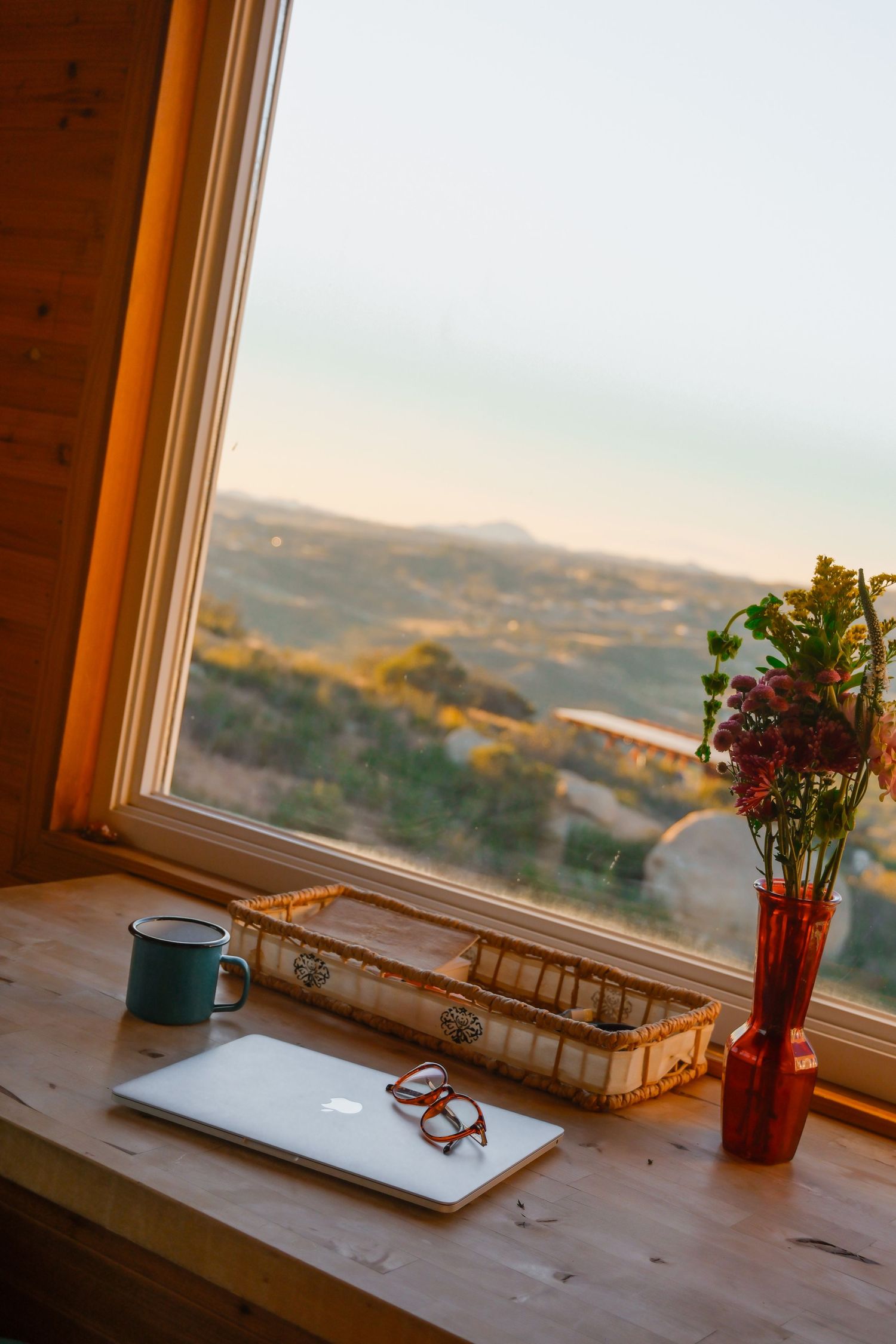 Rustic wooden windowsill workspace with laptop, glasses, mug, and red flower vase overlooking a scenic mountain landscape.