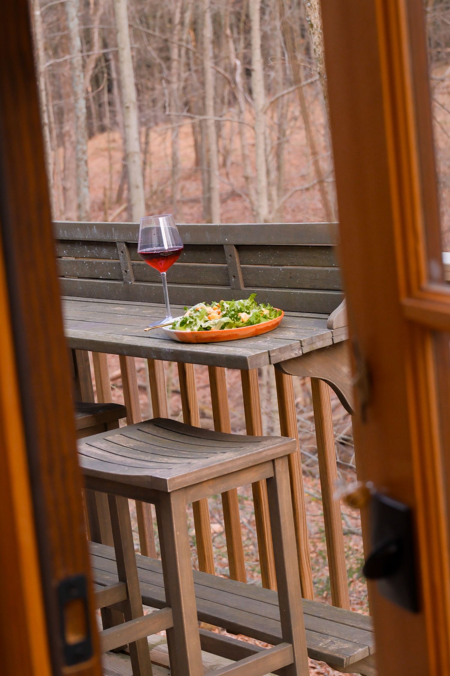 Outdoor wooden deck table set with salad and glass of red wine overlooking autumn forest through open doors.