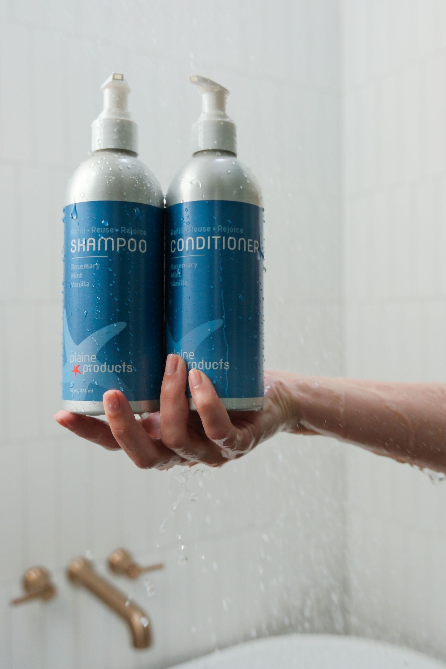 Wet hands holding shampoo and conditioner pump bottles under shower spray in white tiled bathroom.