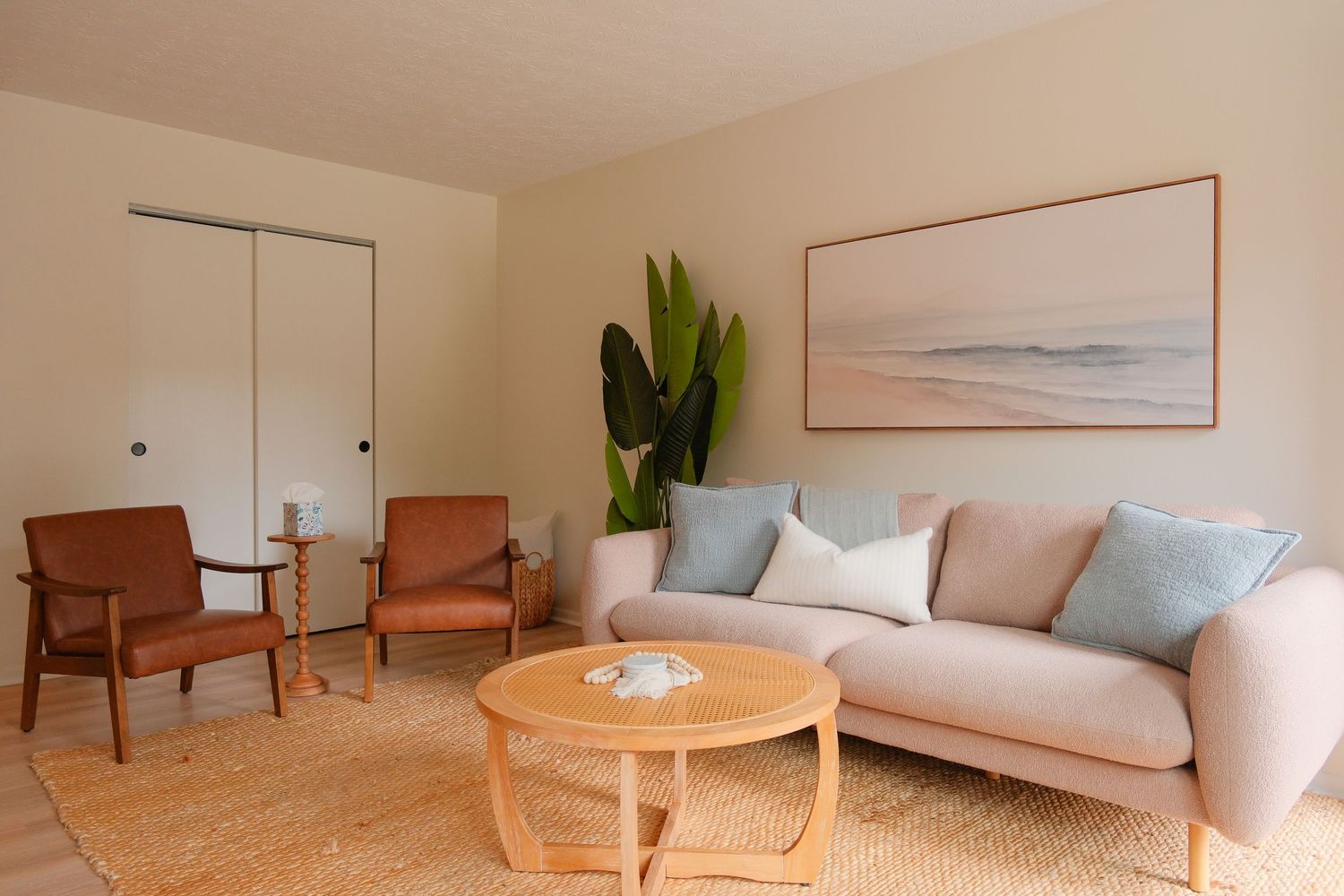 Bright living room with a beige sofa, light blue pillows, round wood coffee table, and two brown leather chairs.