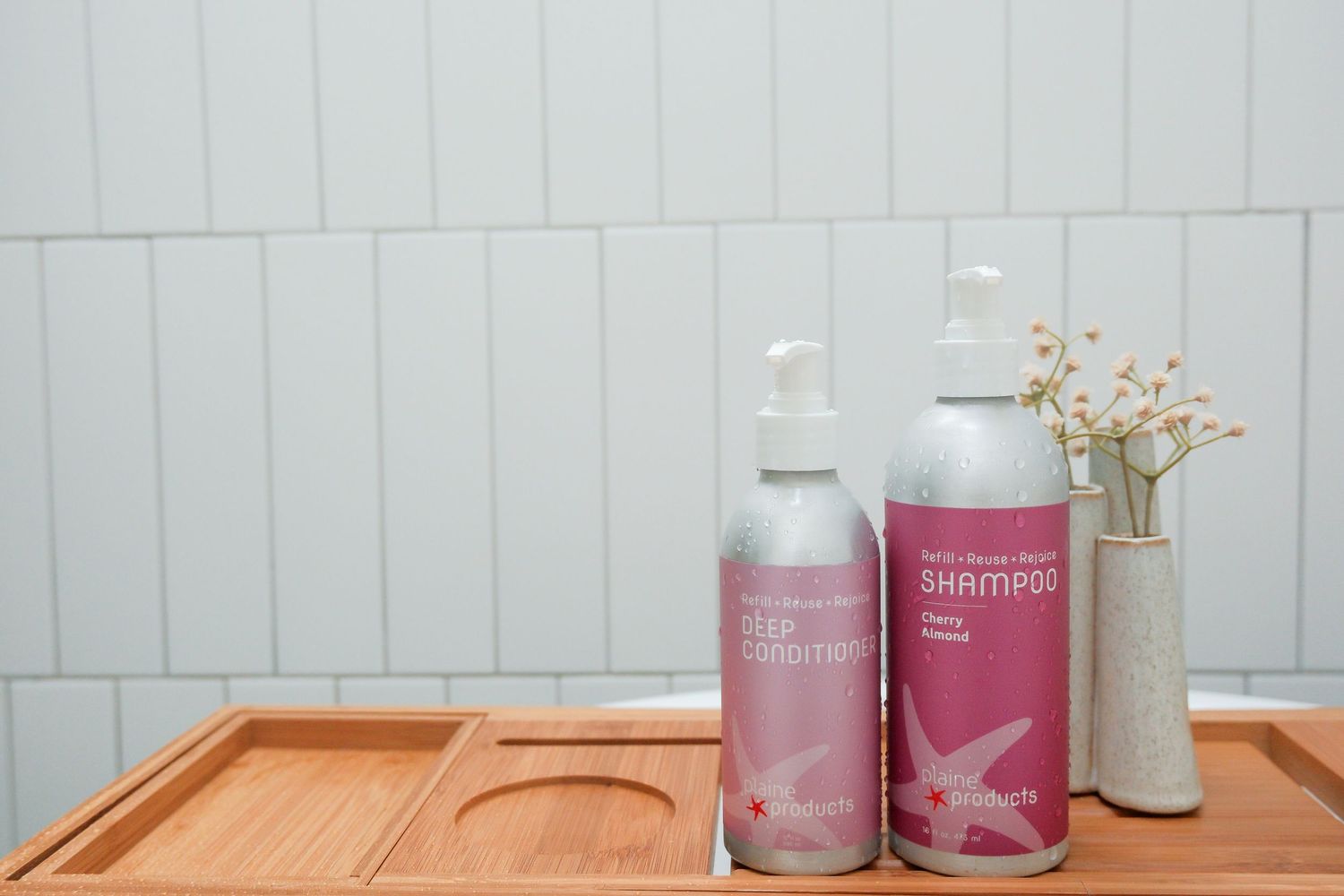 Pink labeled shampoo and conditioner bottles on a wooden tray with a vase against a white tile wall.