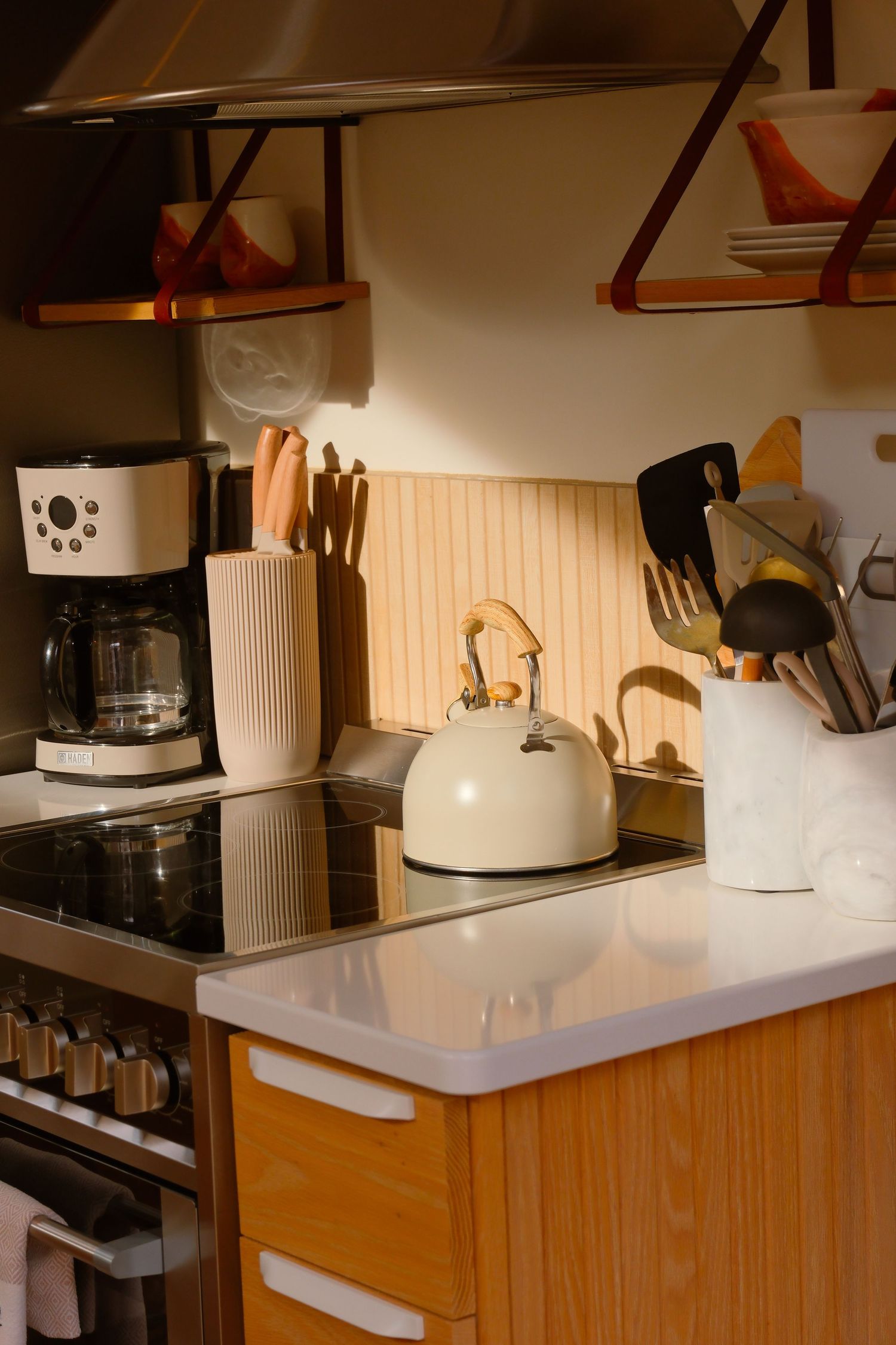 Warm-toned cabin kitchen with cream kettle on induction stove, coffee maker, and wooden shelves with utensils.