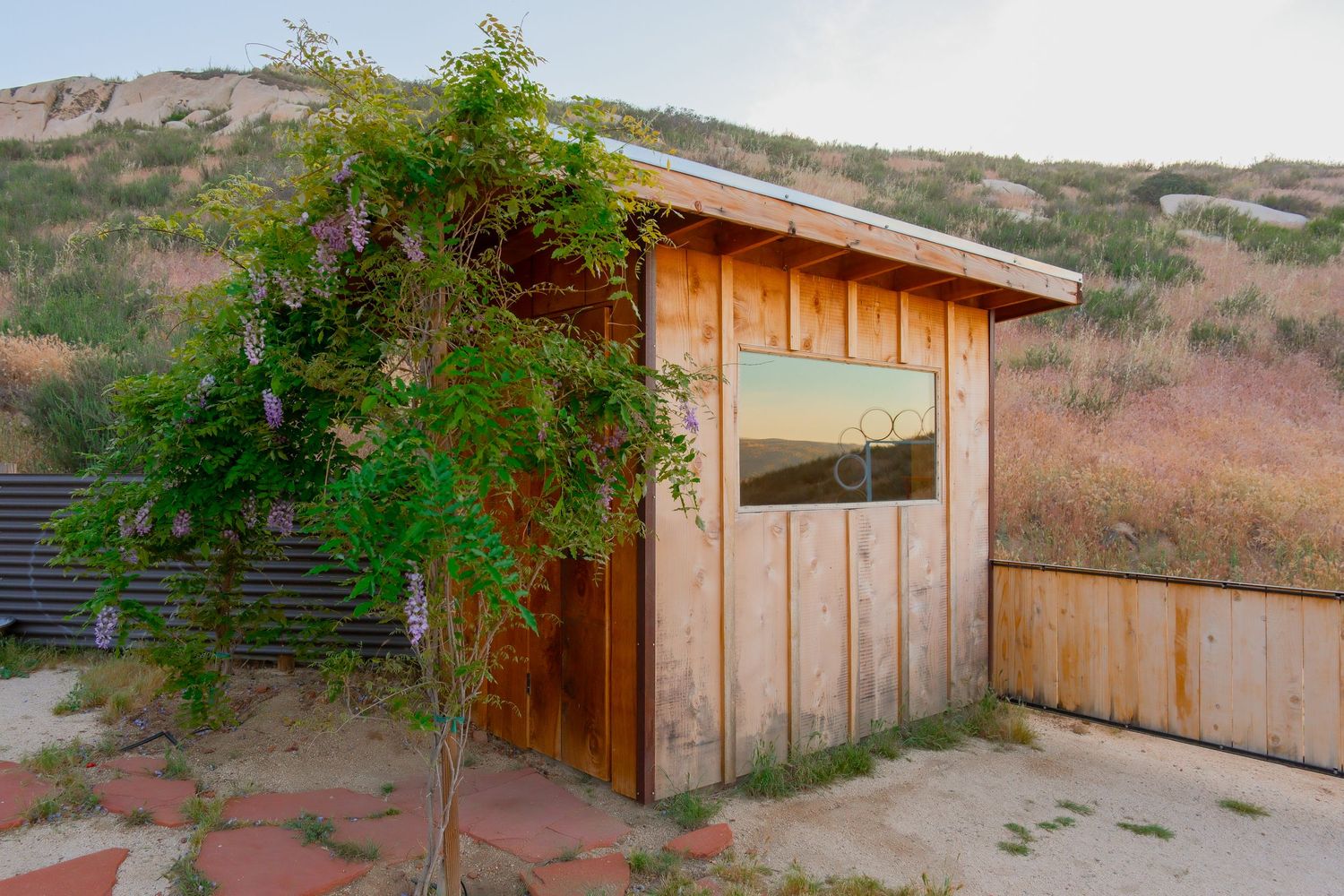 Rustic wooden shed with a small window nestled against a sandy hillside, surrounded by a green tree and dry grass.