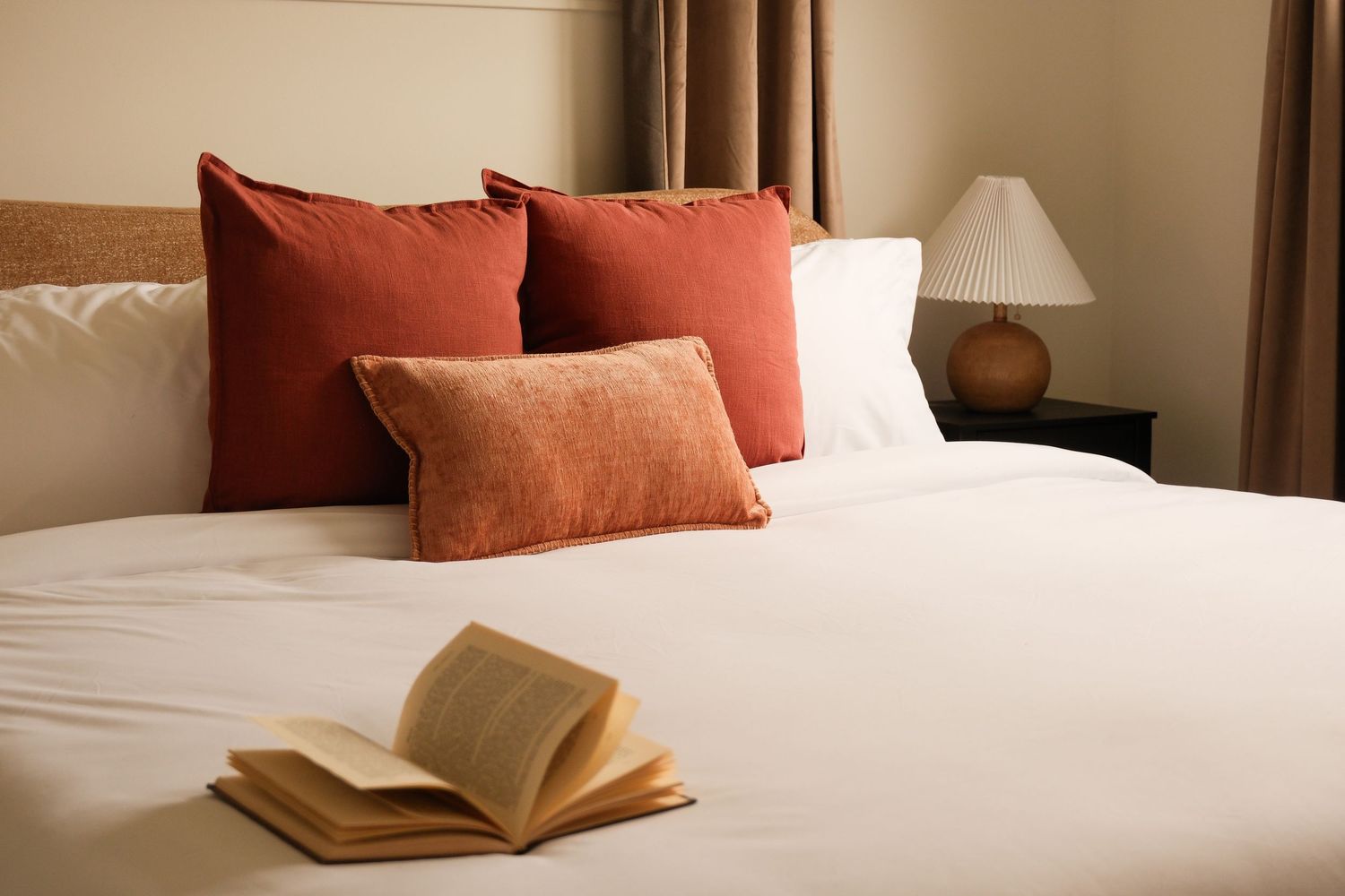 Neatly made bed with red and orange decorative pillows and an open book on white bedding.