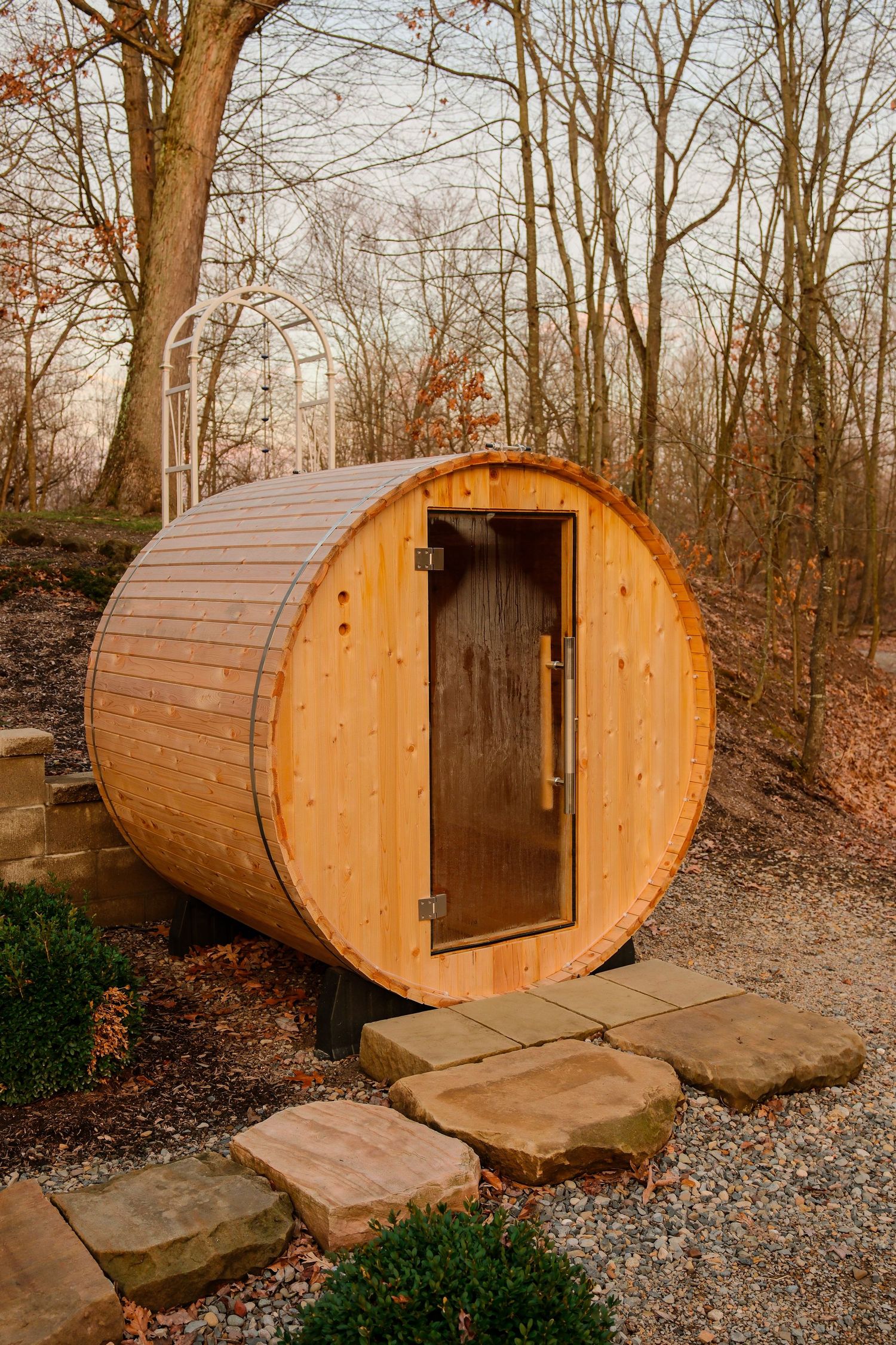 Rustic cedar barrel sauna with glass door, surrounded by stone pathway and bare trees in a serene outdoor woodland setting.