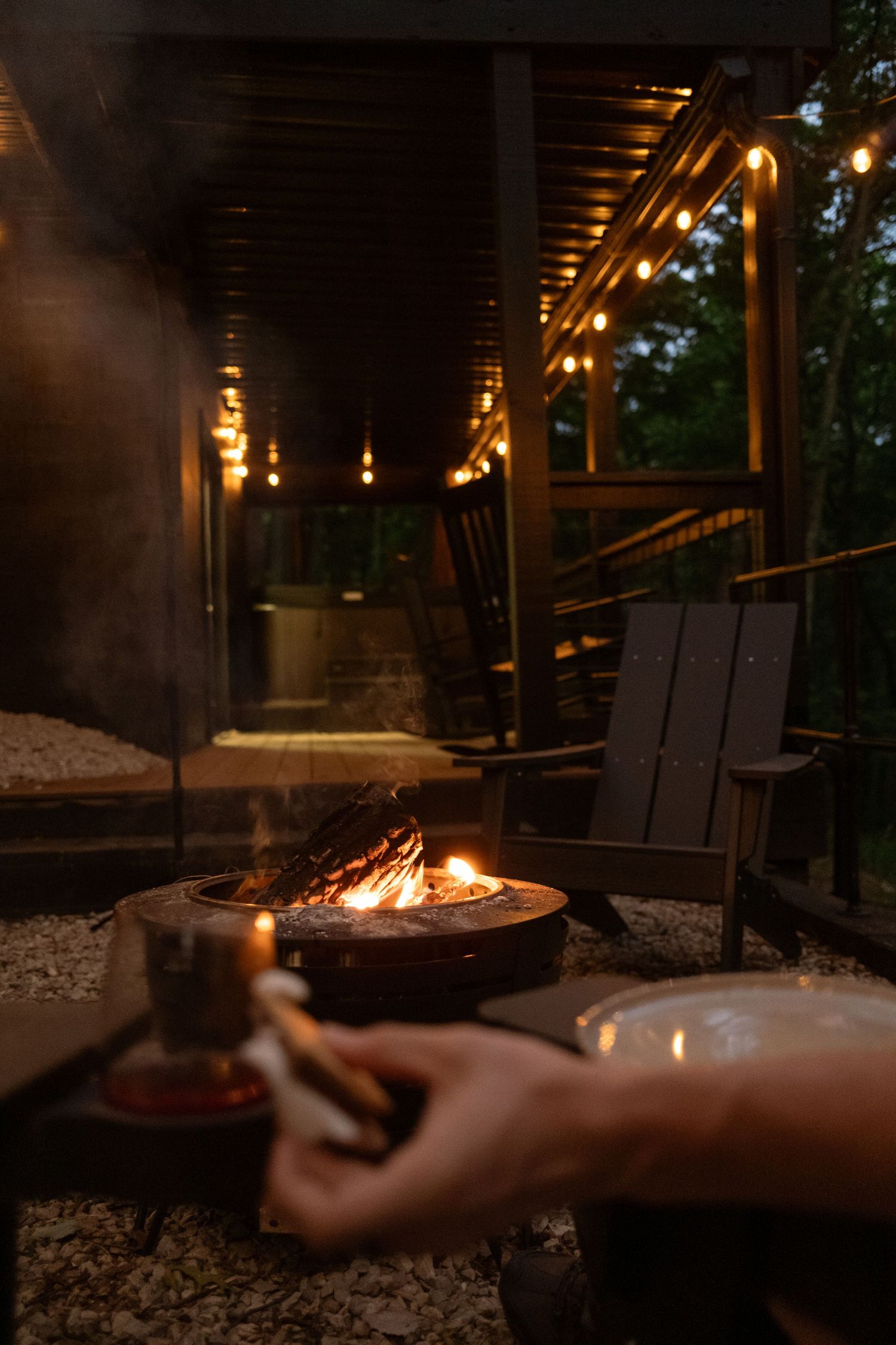 Fire pit glowing on wooden deck at night with string lights overhead.