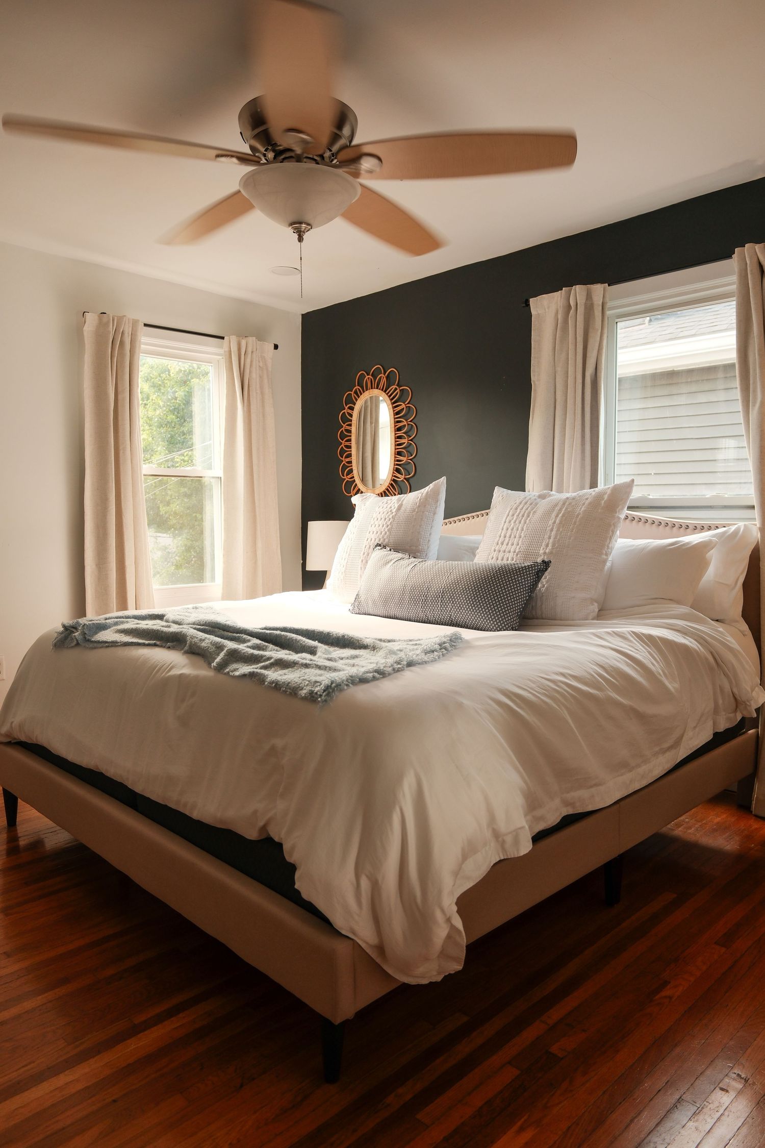 Cozy bedroom with dark accent wall, king bed with white bedding, hardwood floors, ceiling fan, and two windows with cream curtains.