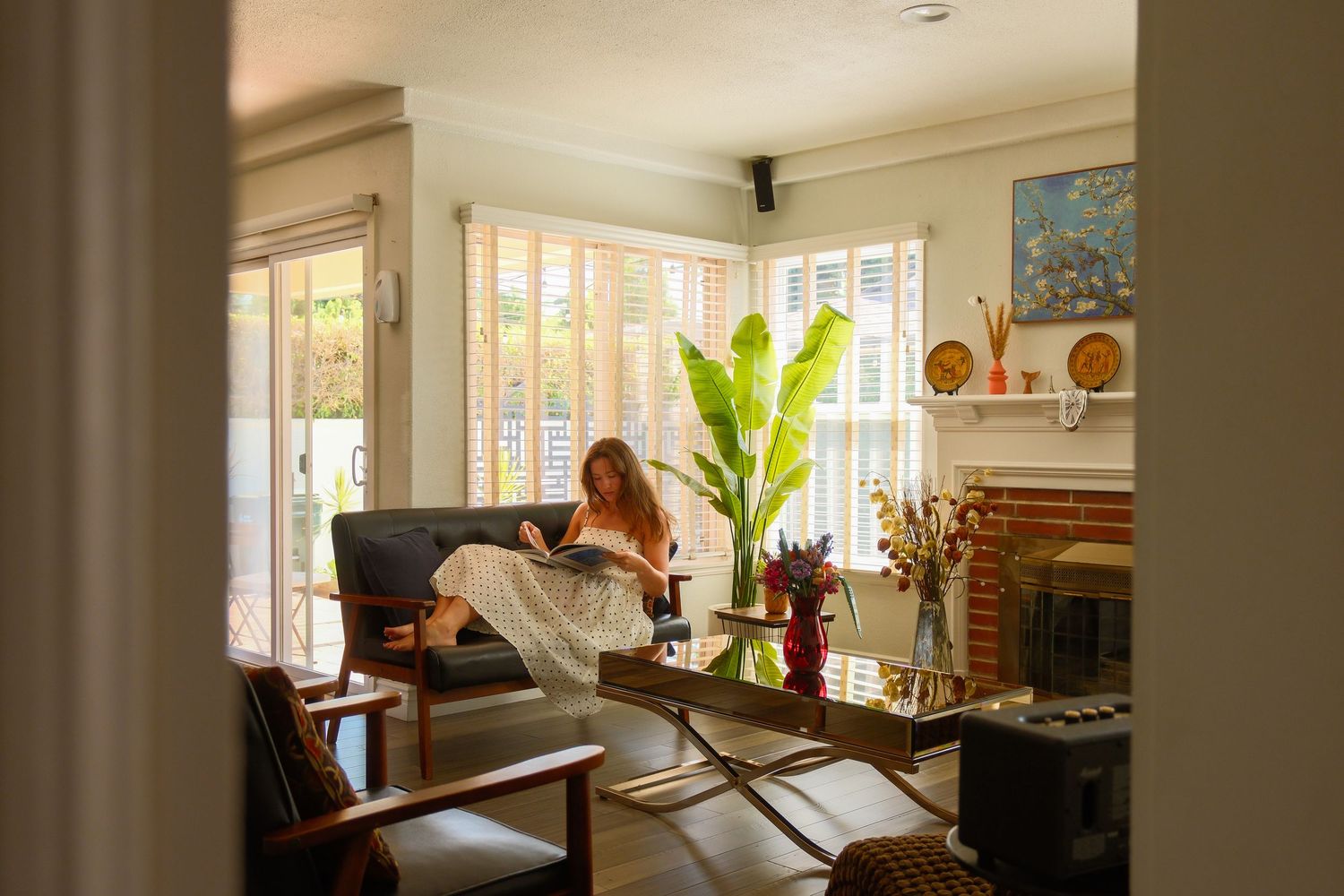 Woman relaxing on sofa in bright living room with large plants, fireplace, and natural light through shuttered windows.