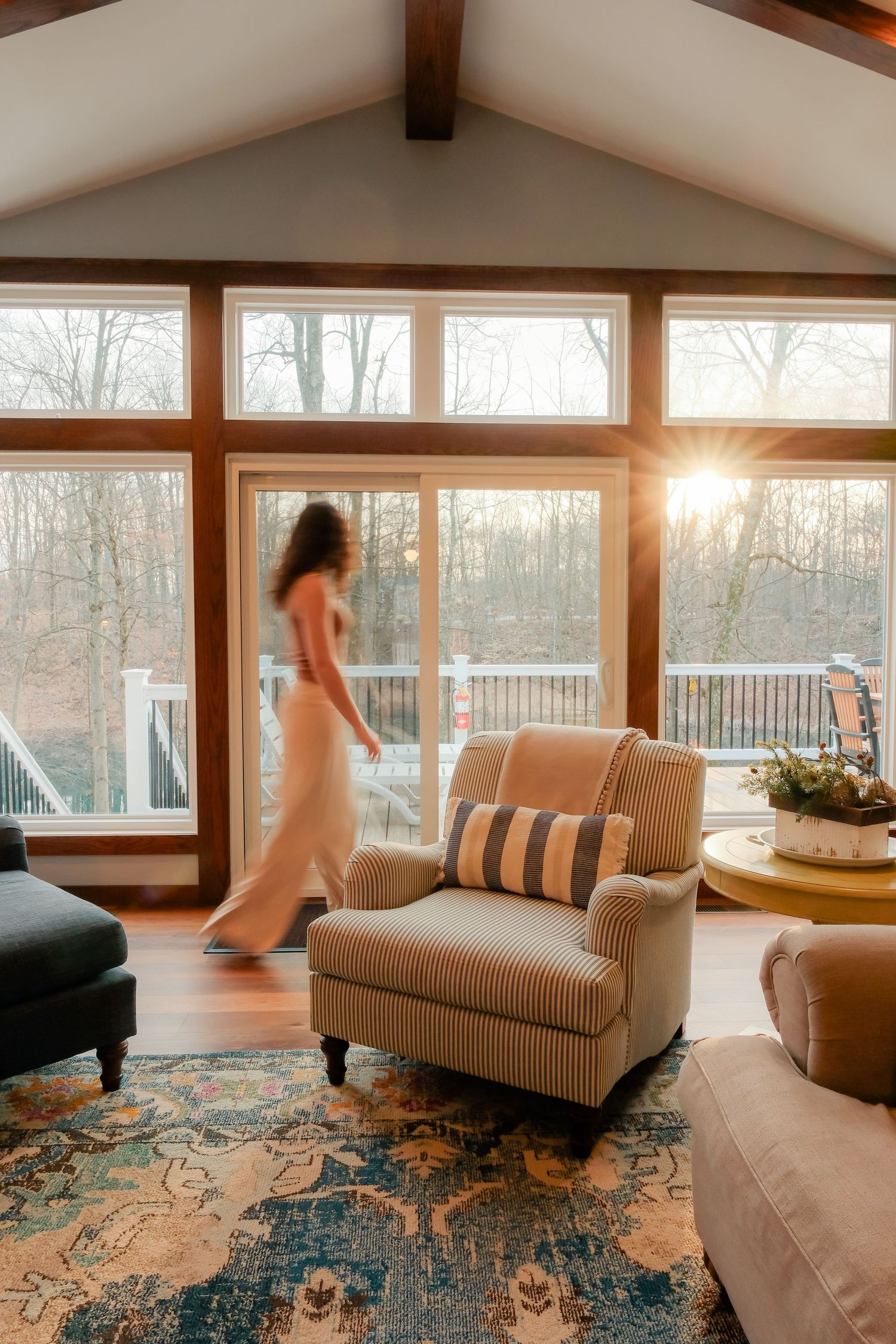 Sunlit living room with comfortable chaise lounges and a blue patterned area rug overlooking wooded views through large windows.