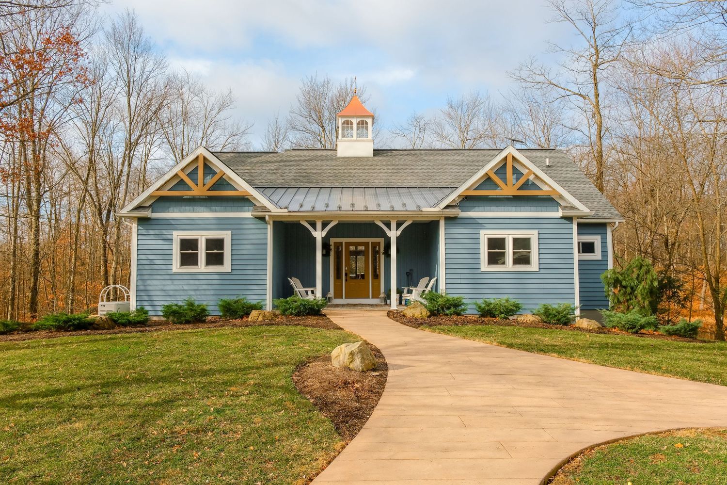 Blue craftsman-style home with wood accents, metal roof, covered porch, and curved concrete driveway surrounded by trees.