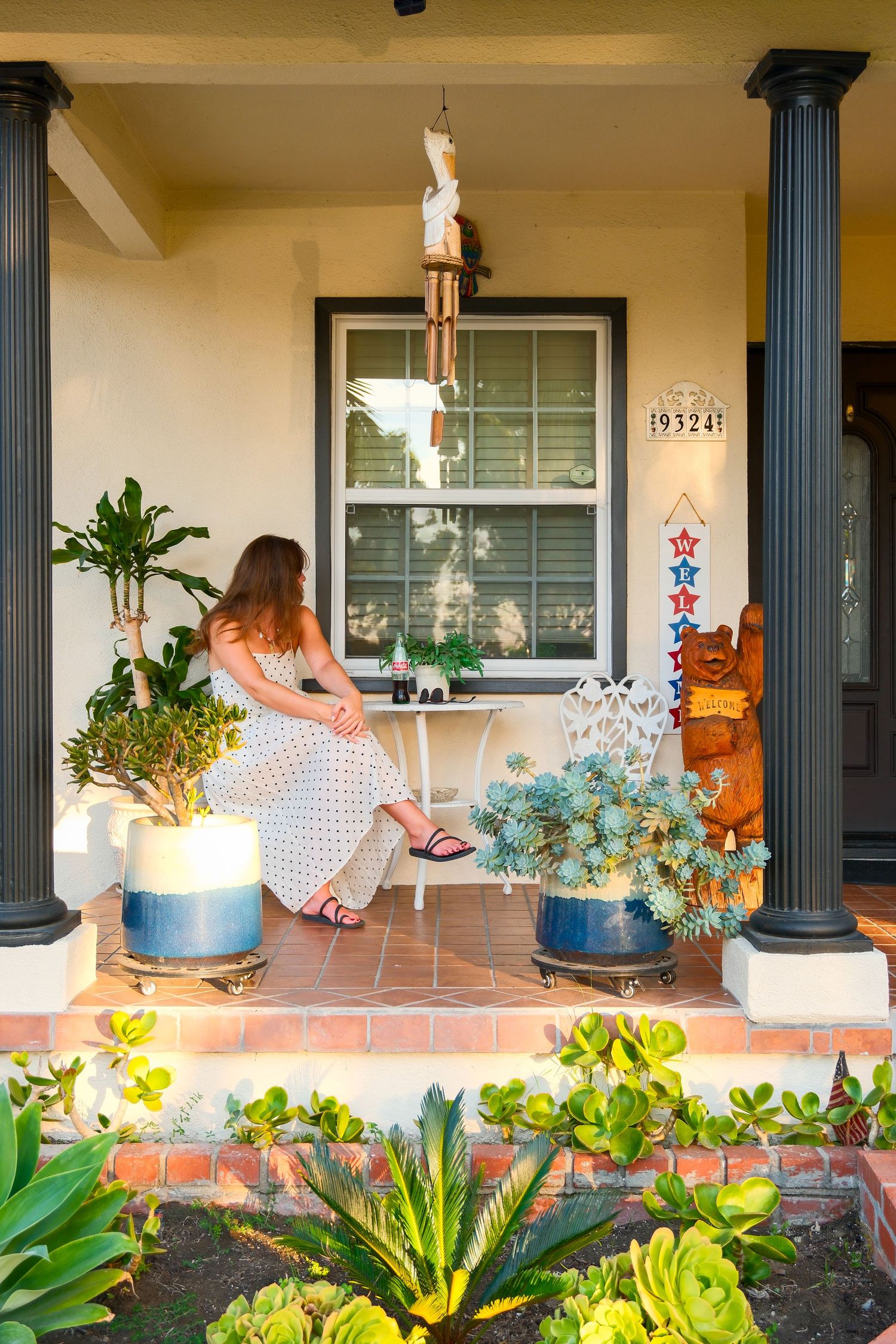 Woman in floral dress sitting on a decorated front porch with succulents, potted plants, and patriotic decor.