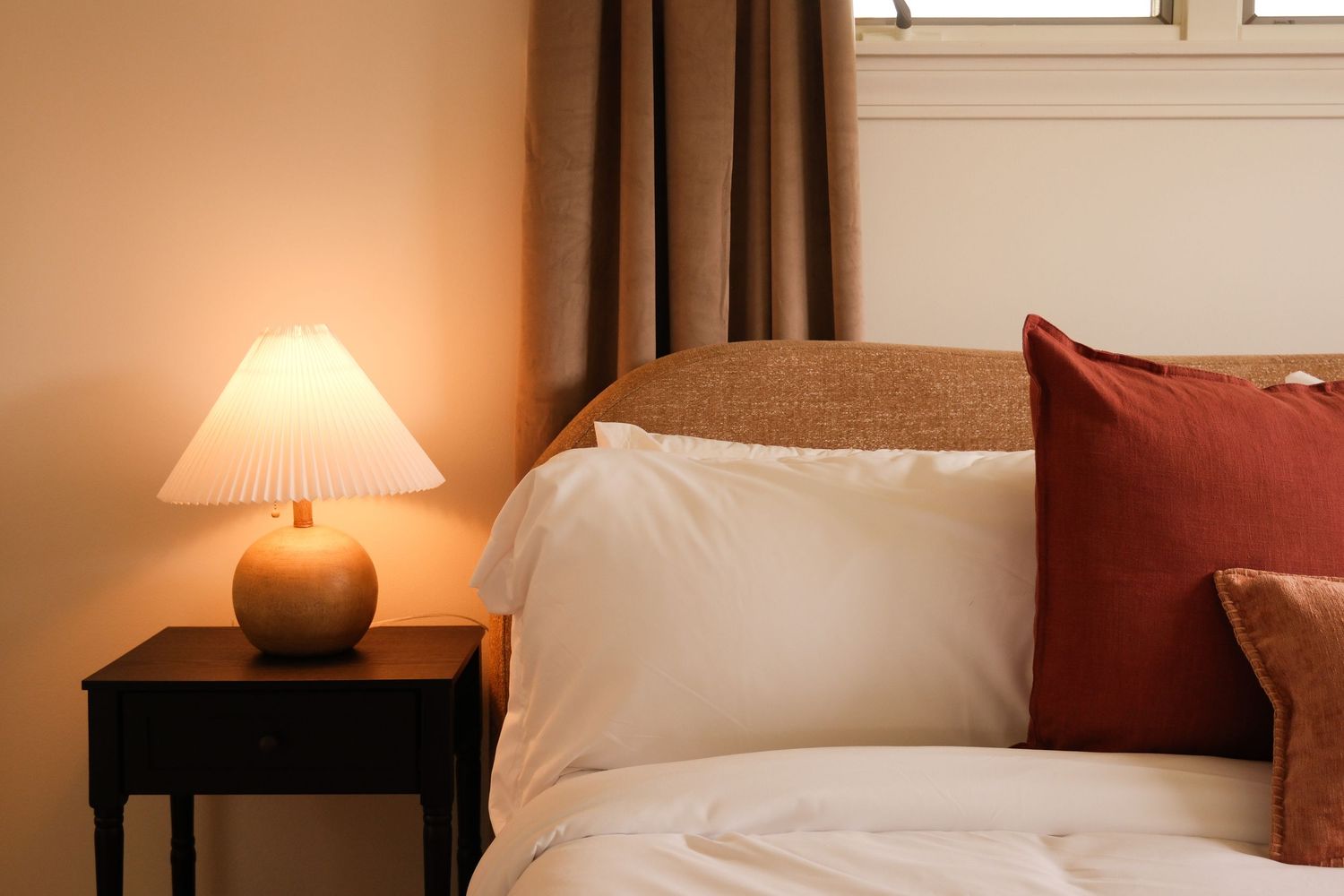 Cozy bedroom with a glowing bedside lamp, white pillows, and red accent cushions on a warm-toned bed.