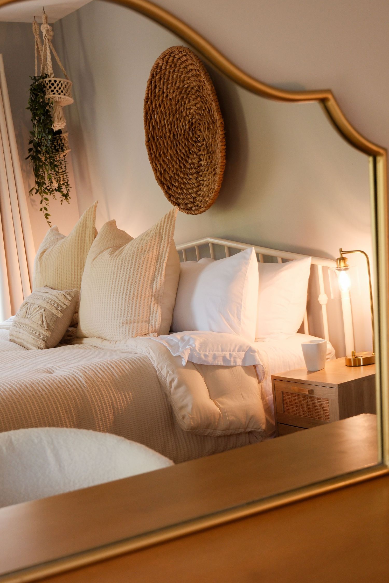 Gold-framed mirror reflecting a cozy bedroom with white pillows, beige bedding, a bedside lamp, and woven wall decor.