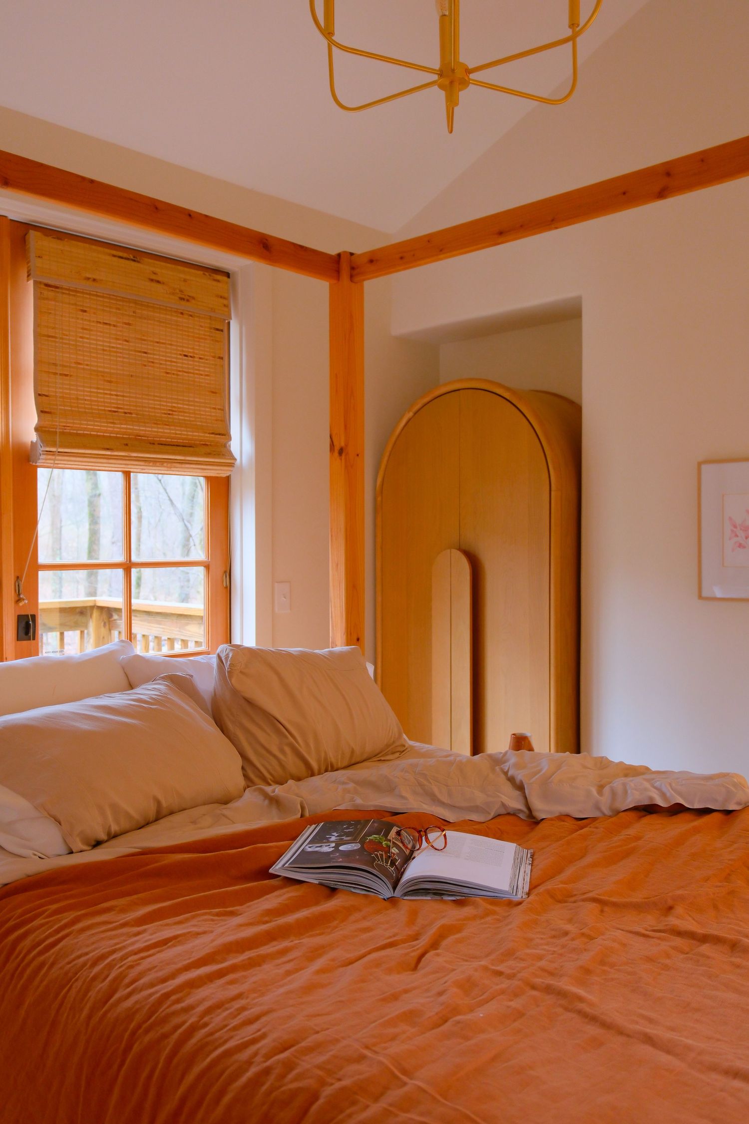 Cozy bedroom with orange linen bedding, wooden accents, bamboo blinds, and an open book on the bed.