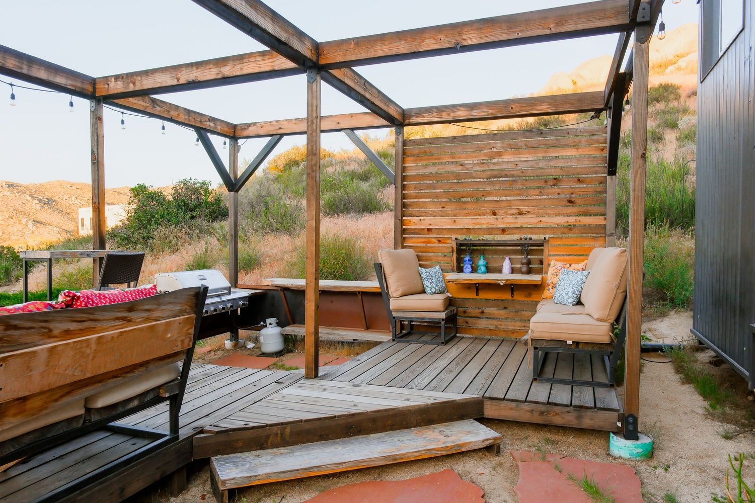 Rustic outdoor deck with wood pergola, cozy seating, string lights, and natural hillside backdrop.