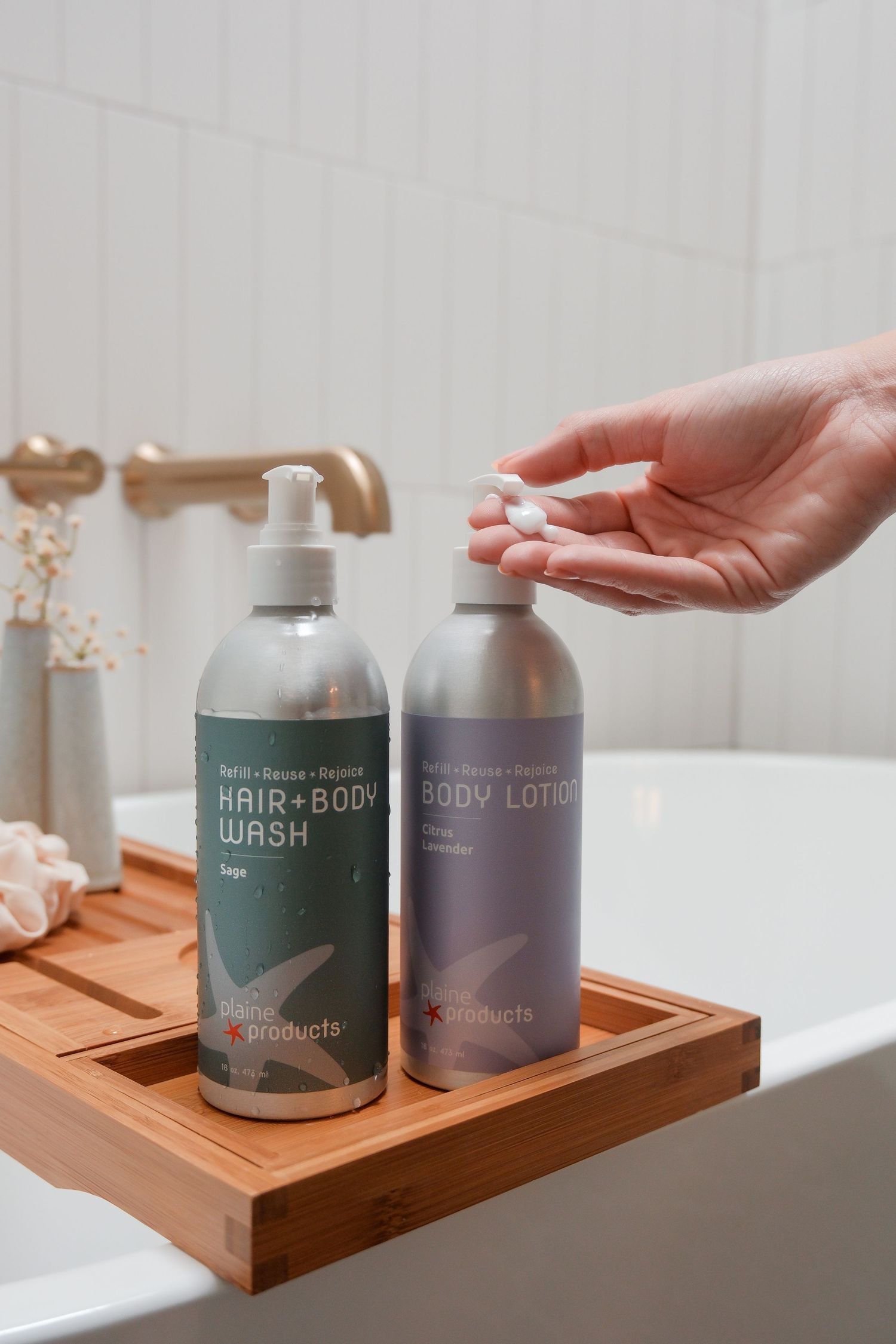 Hand pumping eco-friendly body lotion bottle on bamboo bath tray beside hair and body wash in white tiled bathroom.