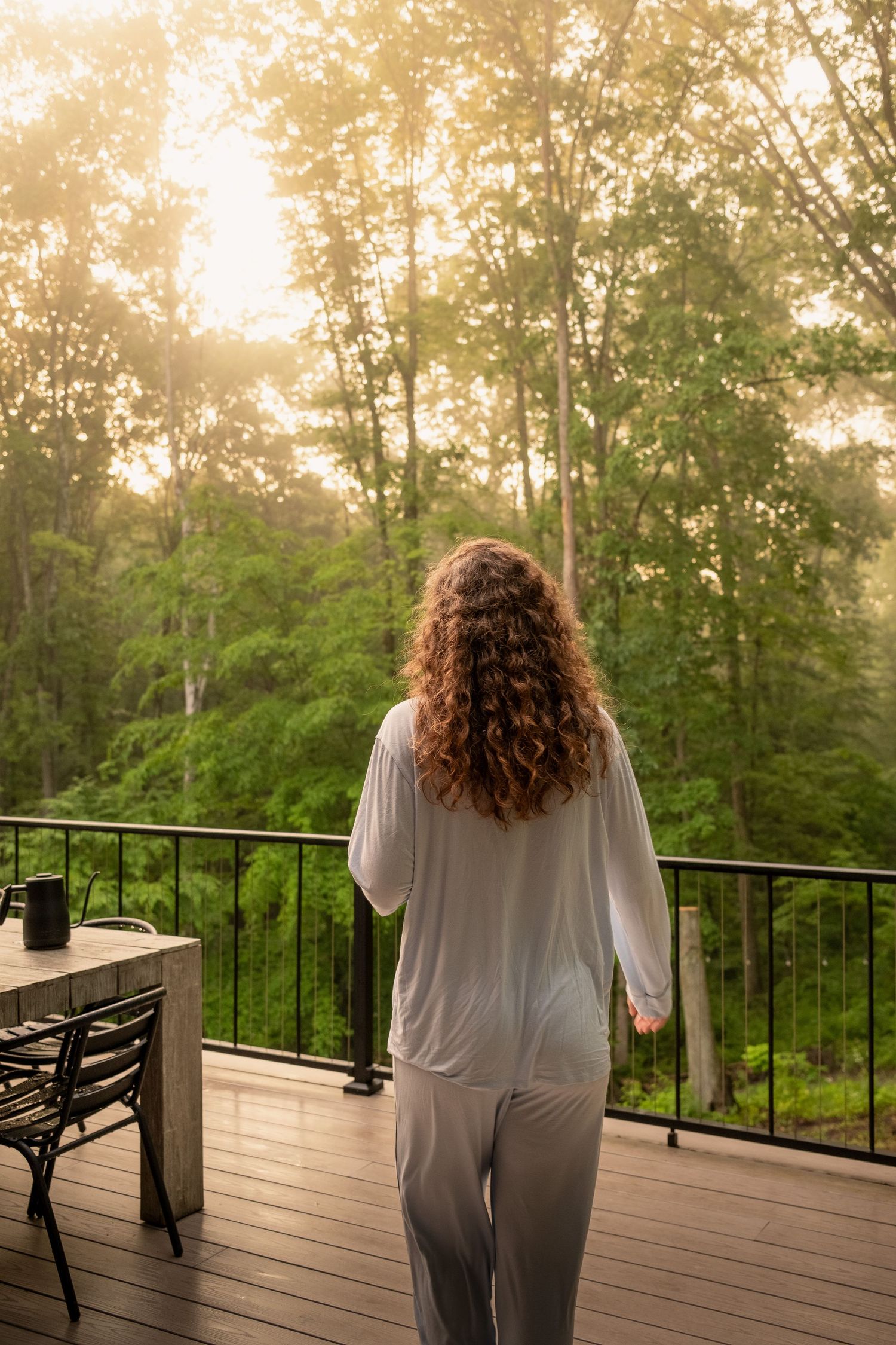 Person in loungewear enjoying morning view from wooden deck surrounded by misty forest landscape.