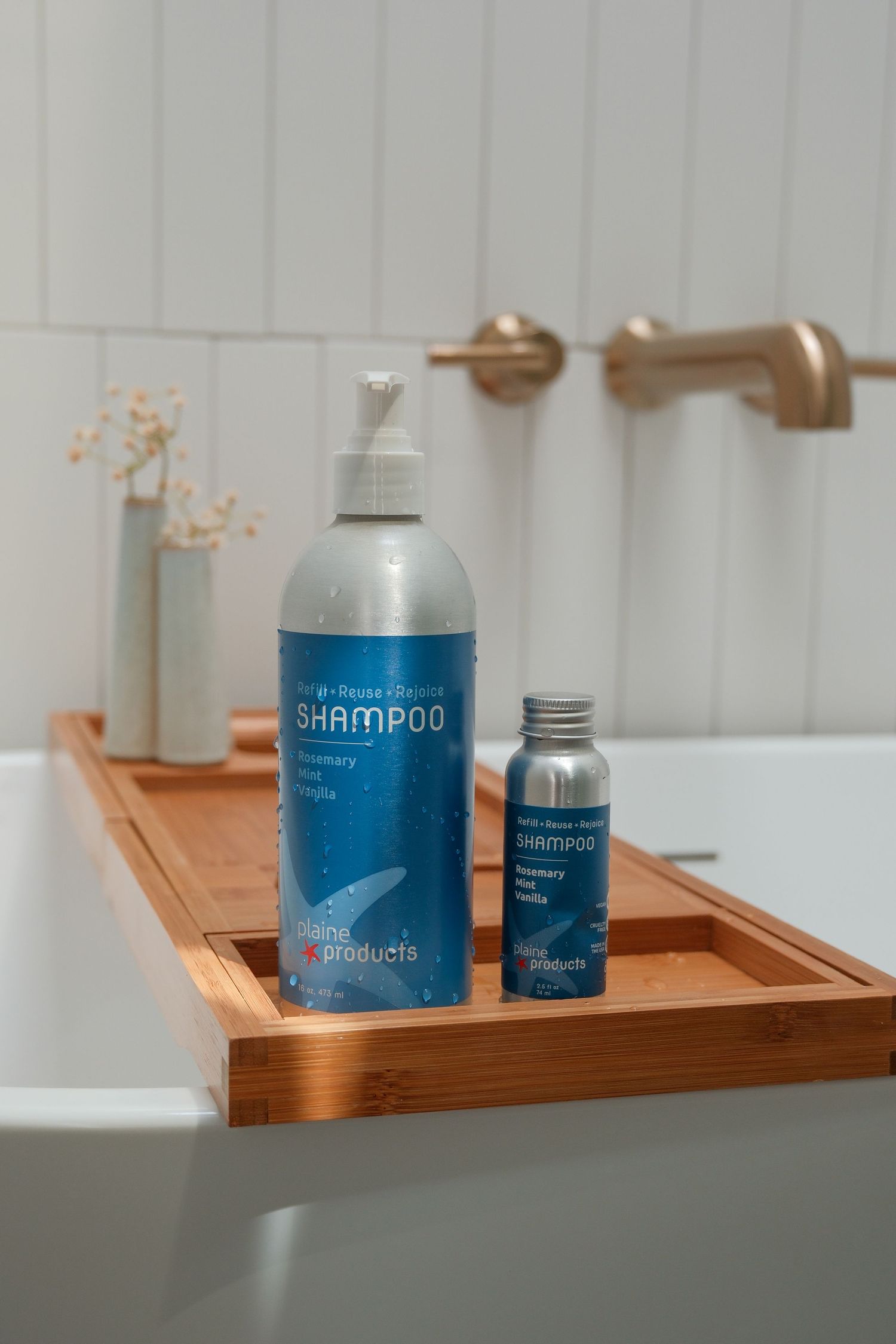 Large and travel size blue shampoo bottles displayed on bamboo bath tray near gold faucet.