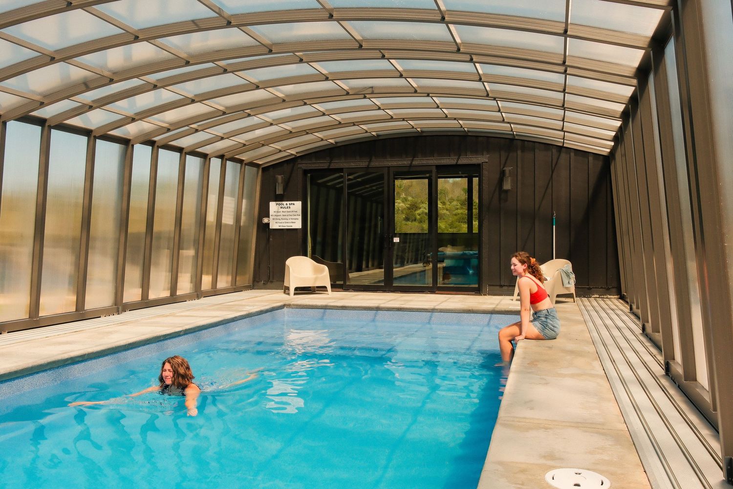 Two women enjoying an indoor swimming pool under a curved glass and metal enclosure with natural light.