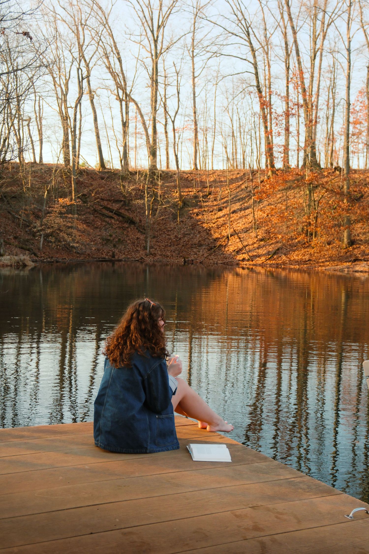 A young woman with curly hair sits on a wooden dock by a calm lake surrounded by autumn trees at golden hour.