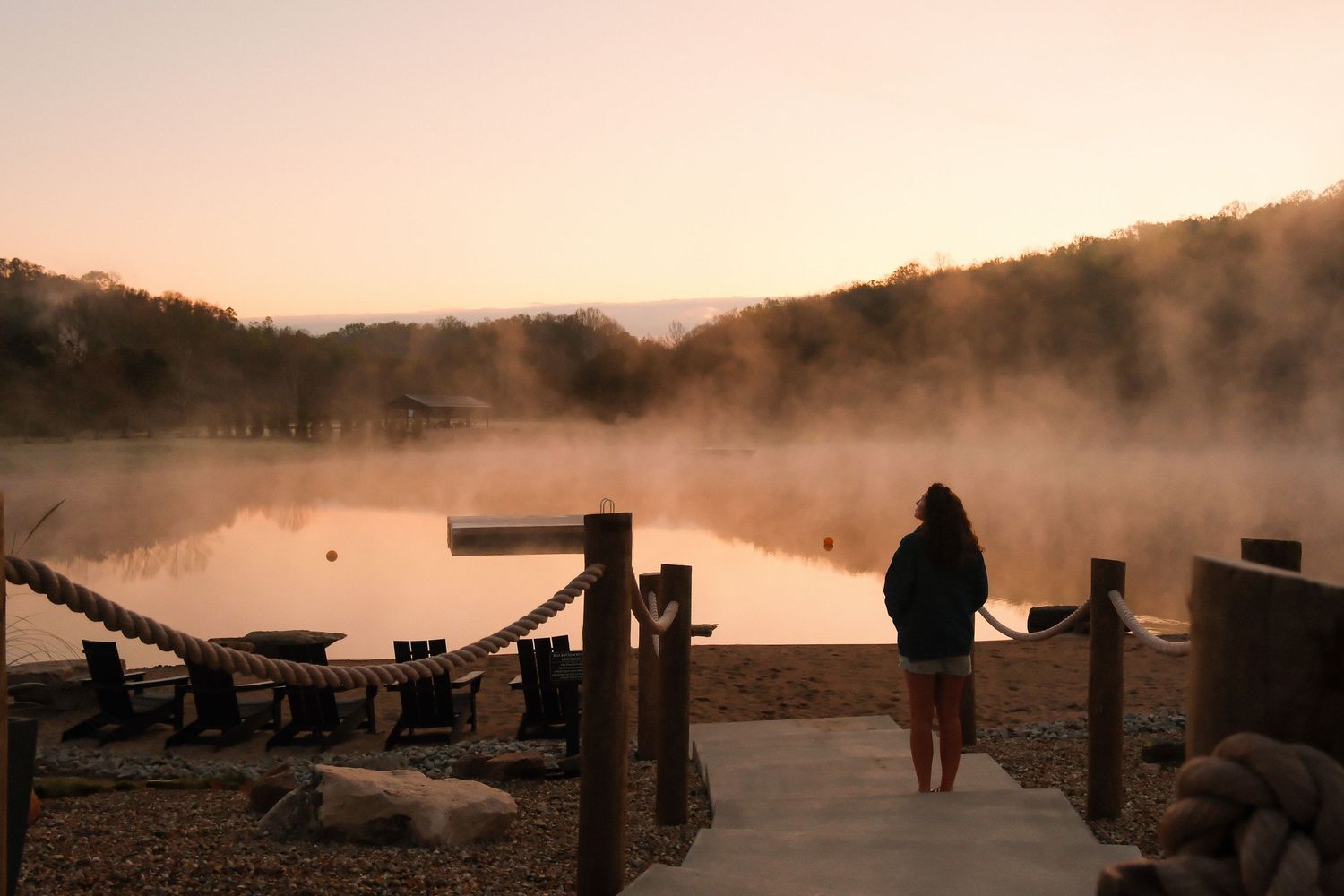 Person standing on a dock at sunrise over a misty lake surrounded by forested hills in warm golden light.