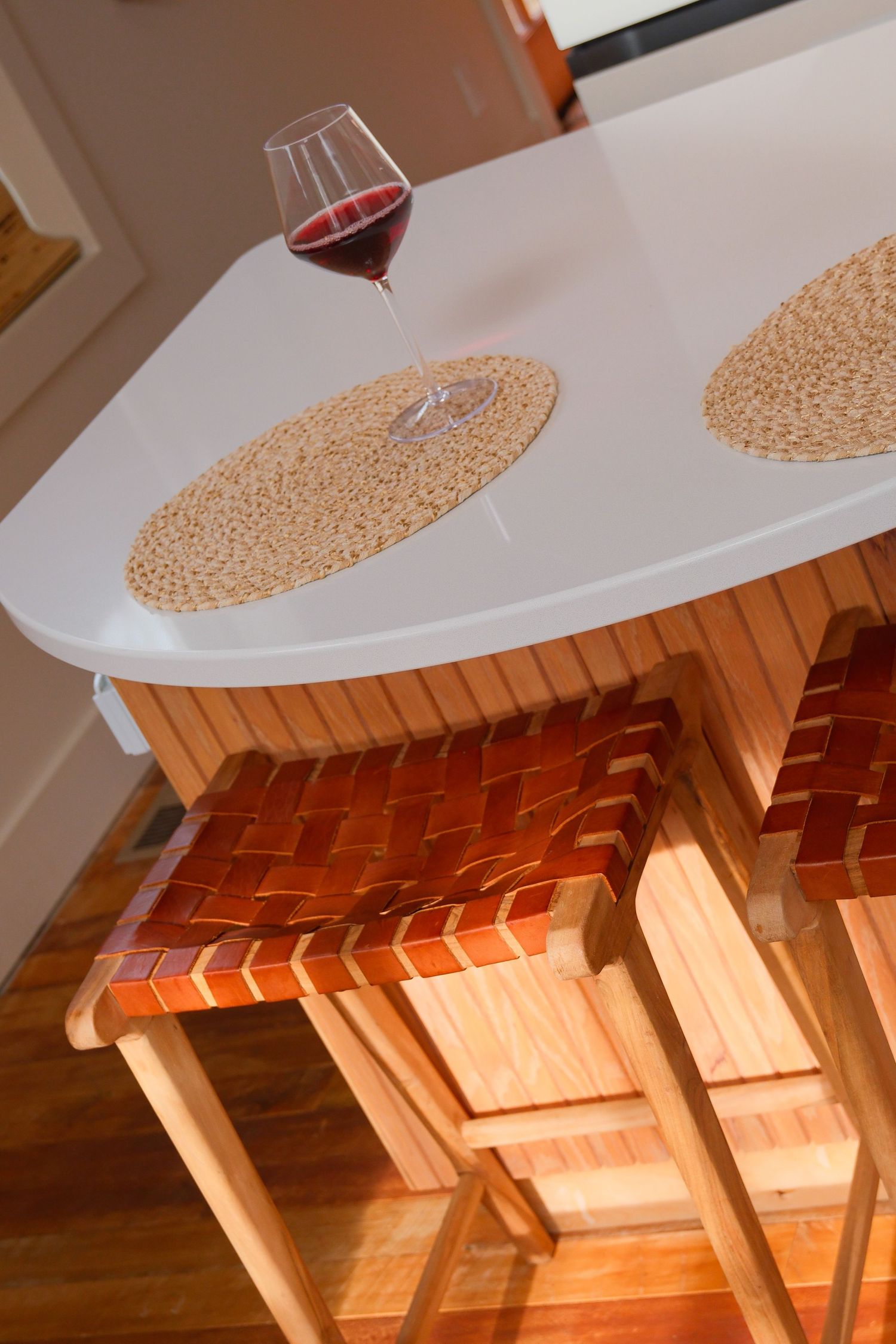 Glass of red wine on a white countertop with woven placemats and leather bar stools below.