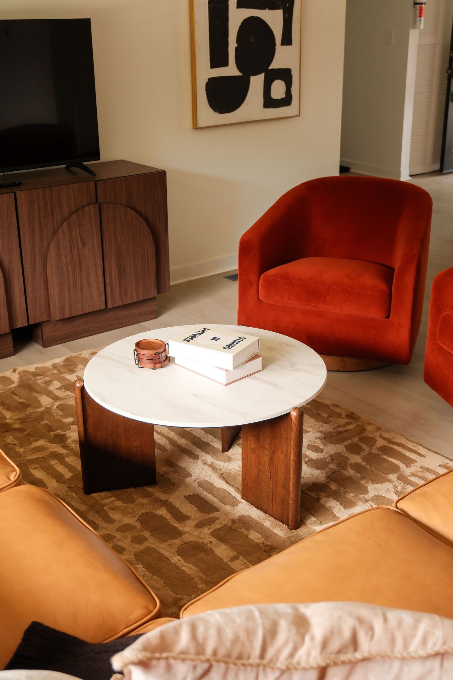 Modern living room with rust velvet armchair, marble coffee table, walnut sideboard, and abstract wall art on light wood floors.