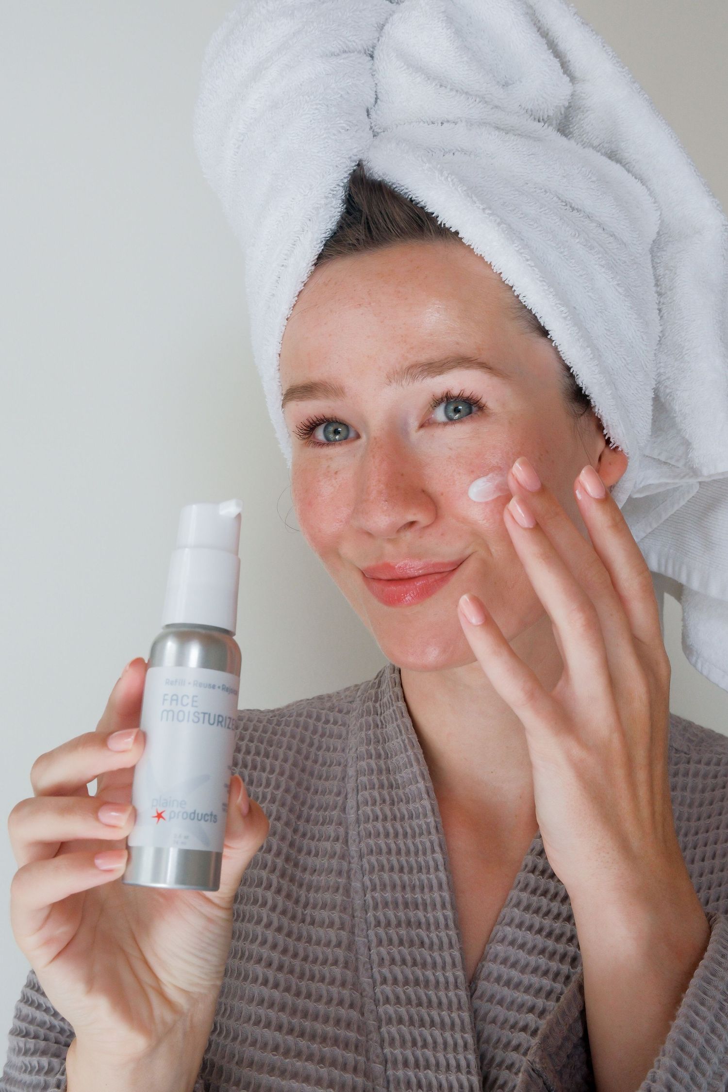 Woman in gray robe and towel turban holding silver face moisturizer bottle while applying cream to cheek.