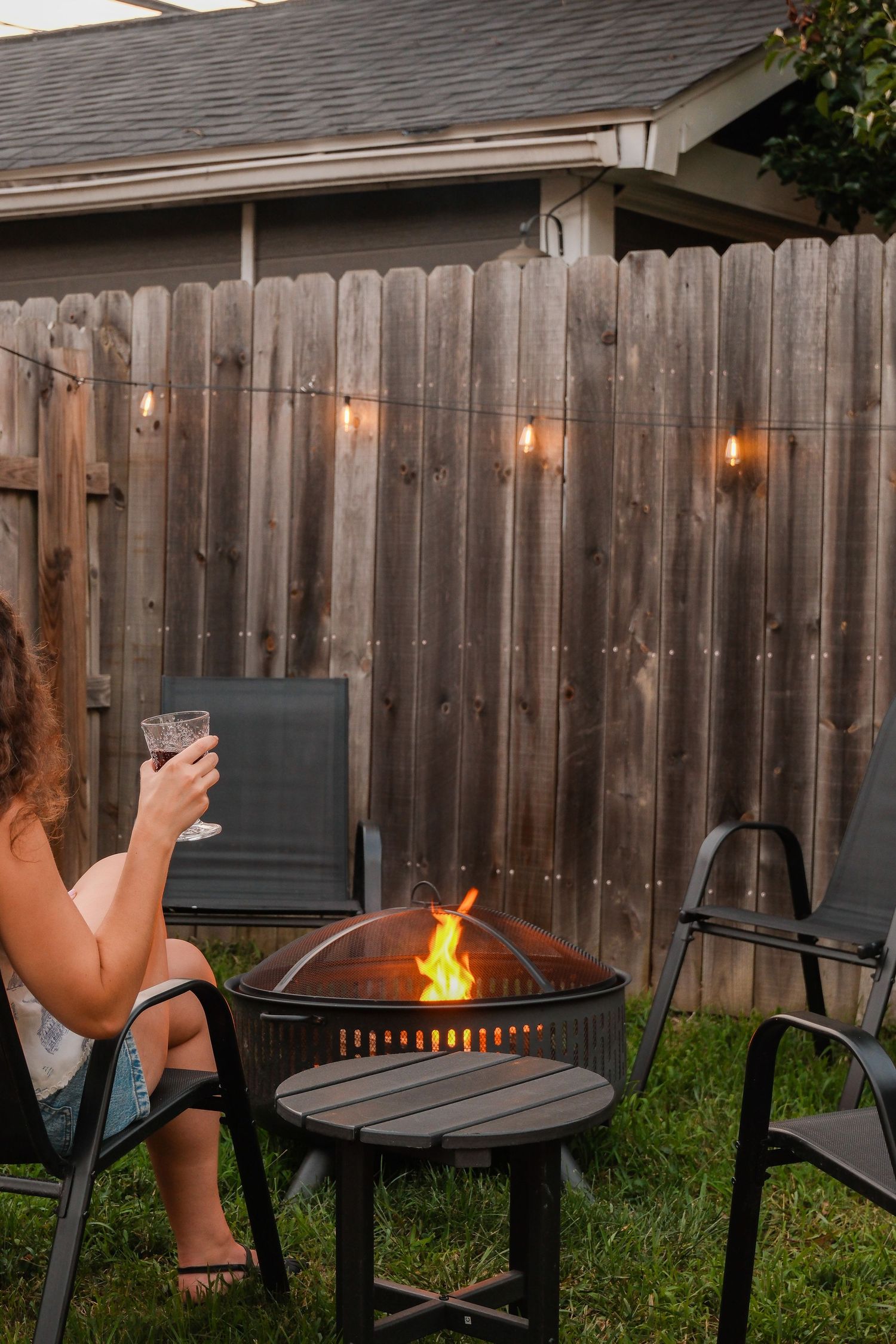 Woman relaxing by an outdoor fire pit with string lights on a wooden fence in a backyard at dusk.