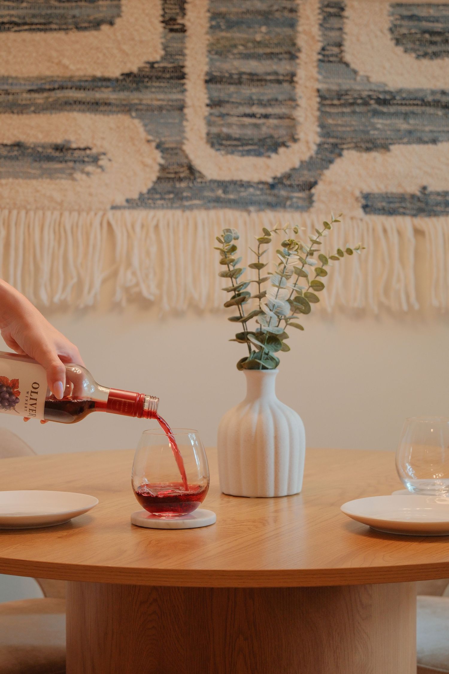 Hand pouring red wine into a glass on a round wooden table with a white vase, eucalyptus, and a woven wall tapestry.