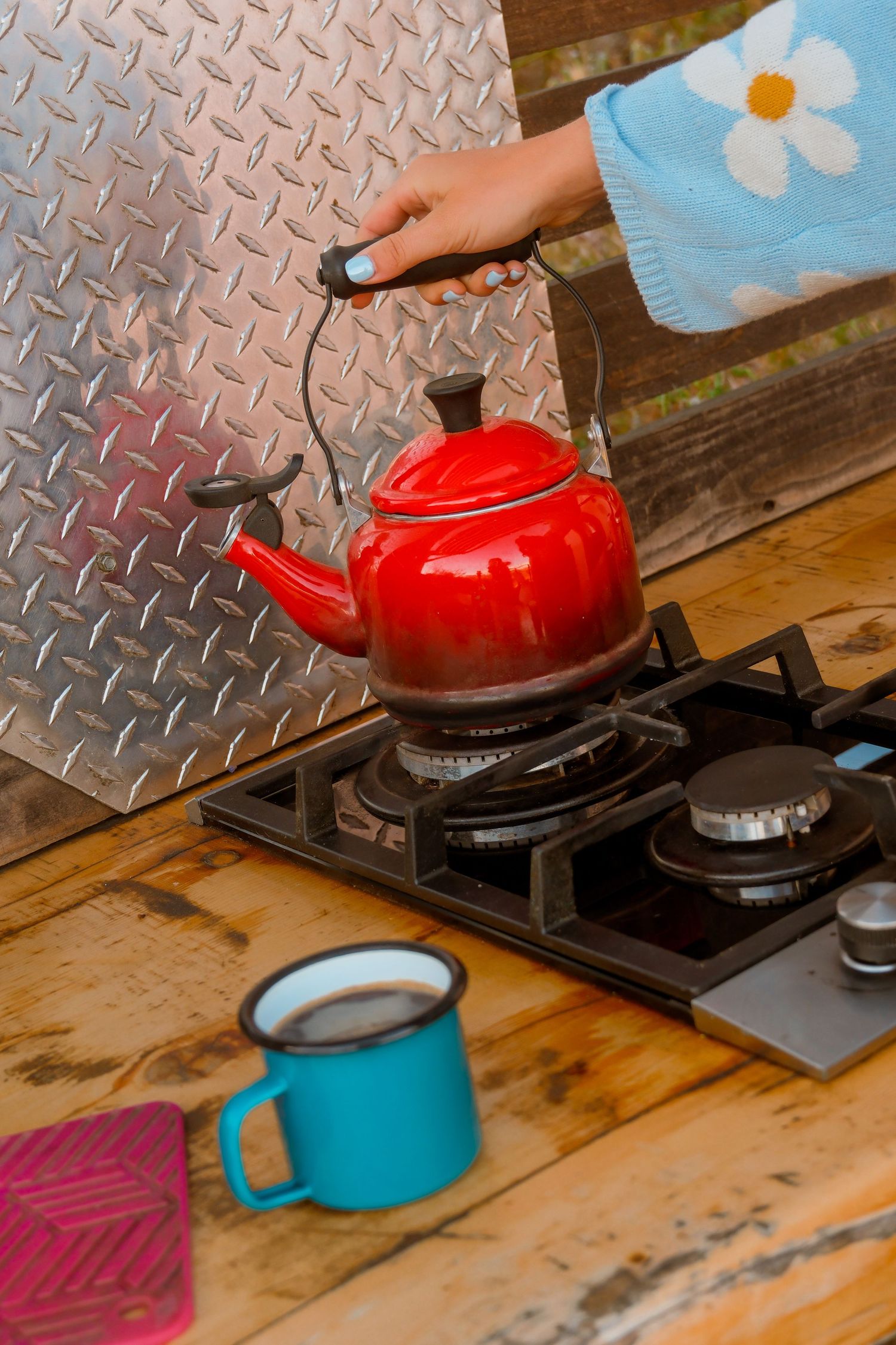 Woman in blue floral sweater placing red kettle on gas stove next to blue enamel mug on rustic wooden counter.