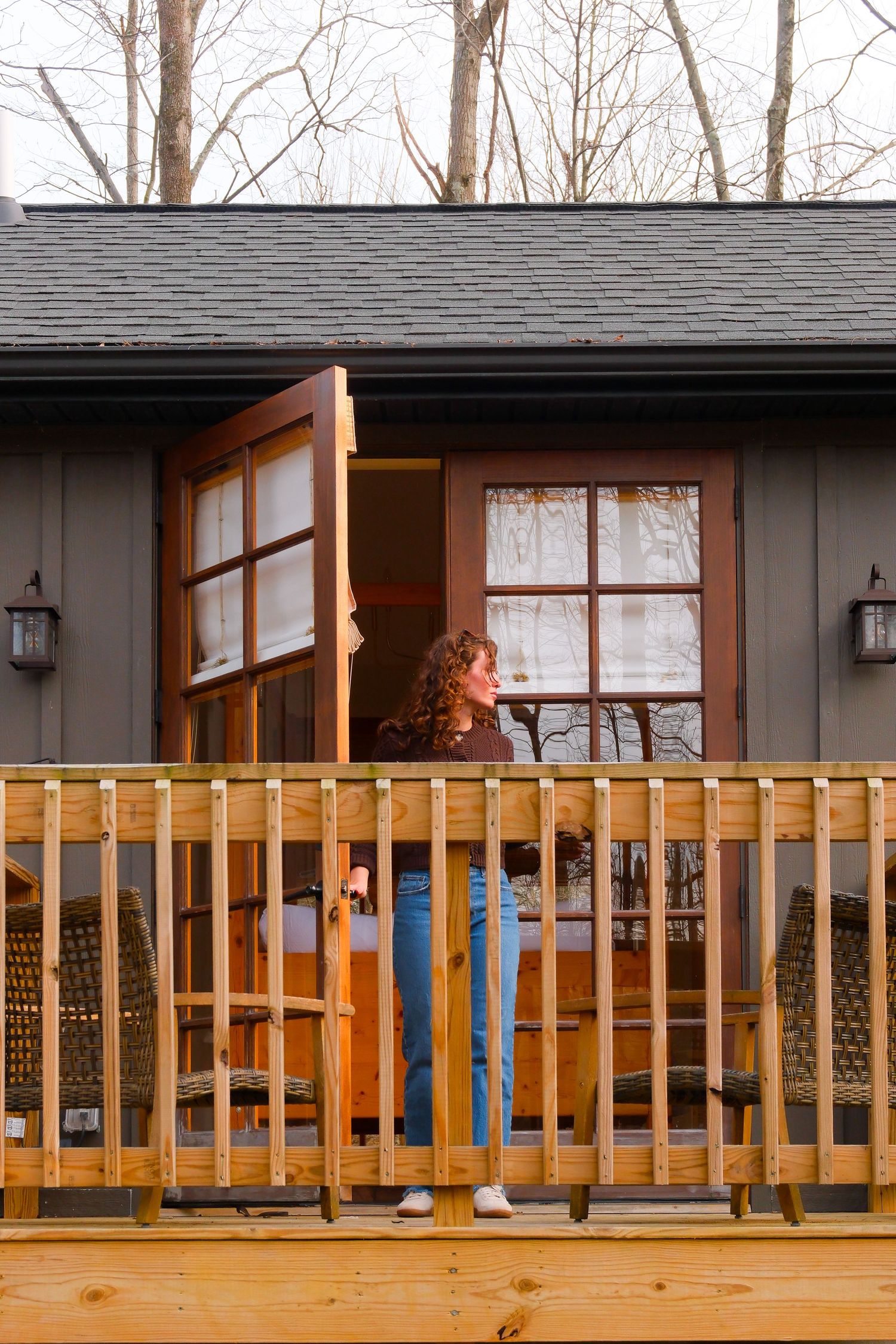 Woman standing on a wooden deck outside a modern cabin with French doors and bare winter trees.