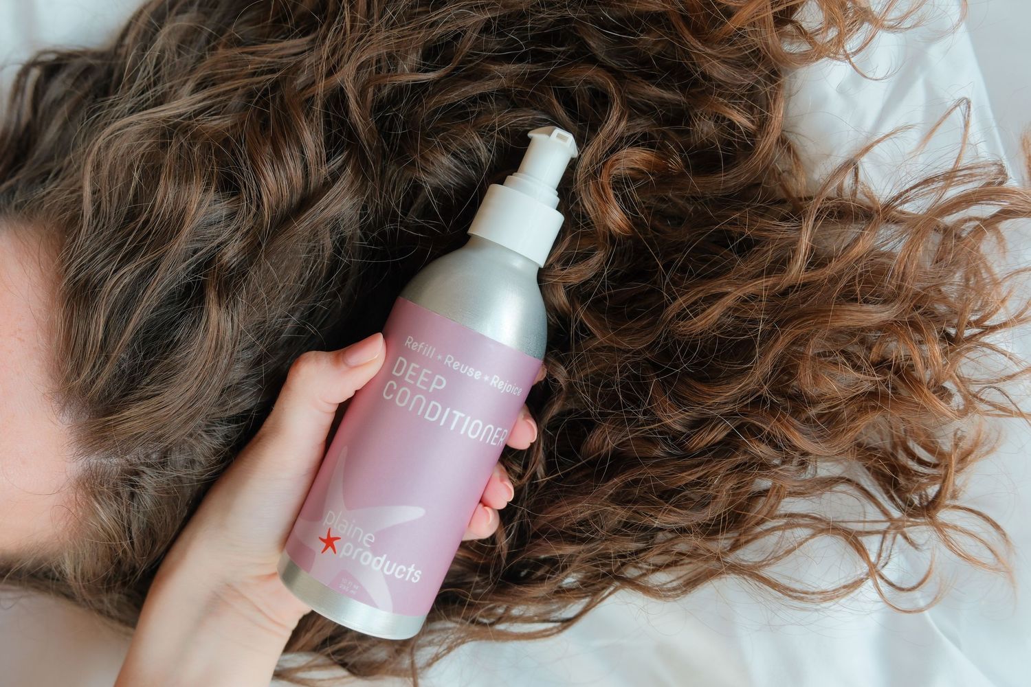 Hand holding a pink hair deep conditioning spray bottle over long, curly brown hair on a white surface.