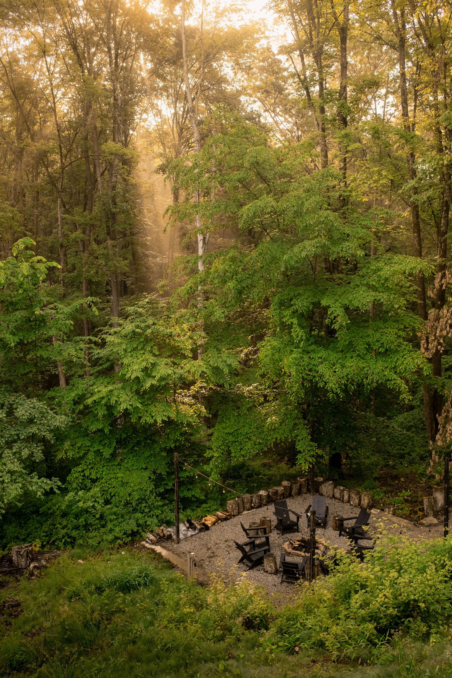 Aerial view of circular fire pit surrounded by Adirondack chairs in wooded setting at sunset.