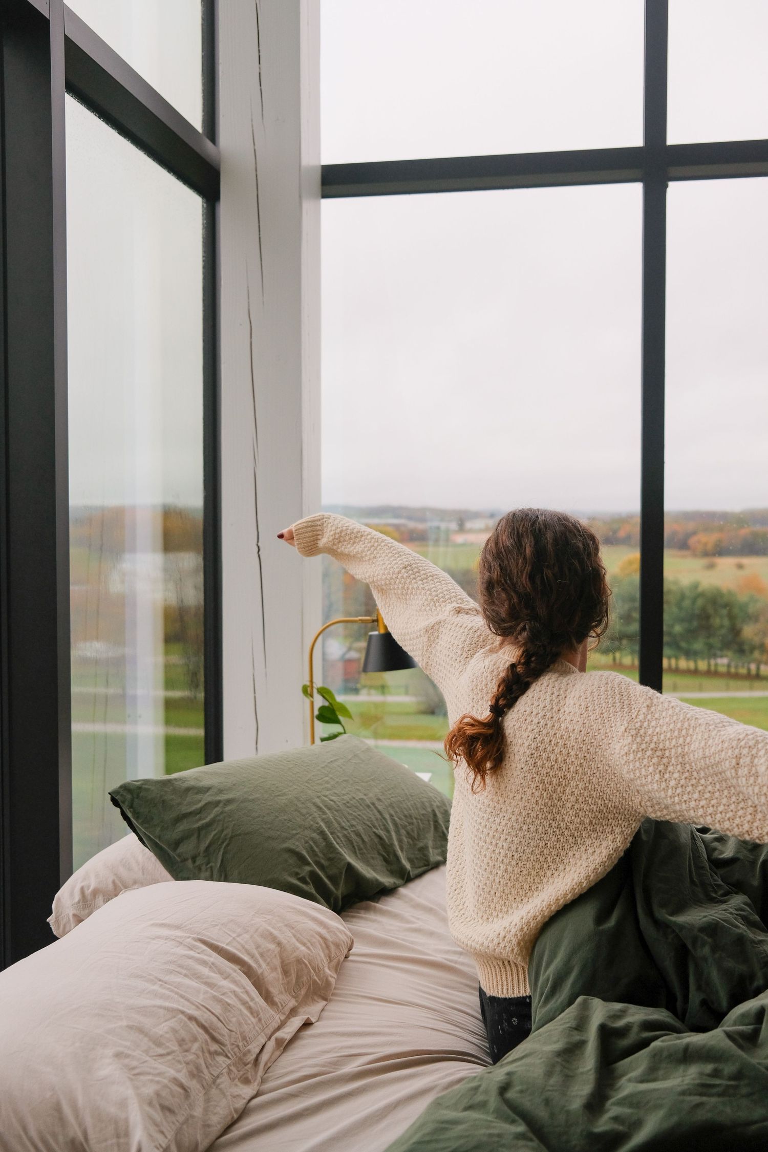 Figure in beige sweater reaching to open window curtains in a bright bedroom with pastoral views.
