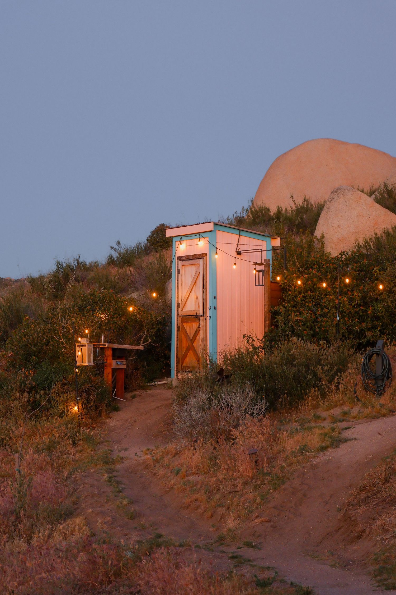 A charming pink outhouse with string lights glows at dusk in a desert landscape with boulders and shrubs.