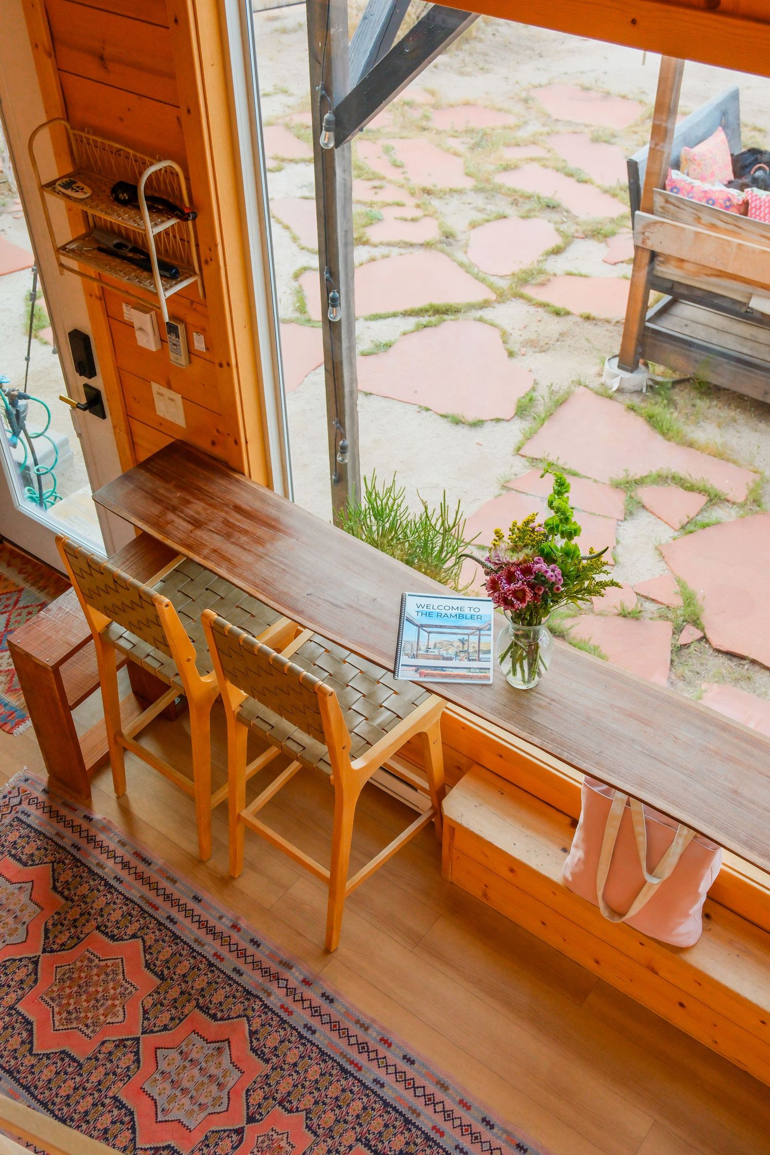 Wooden bar counter with chairs, fresh flowers, and a book beside a large window overlooking a stone patio.