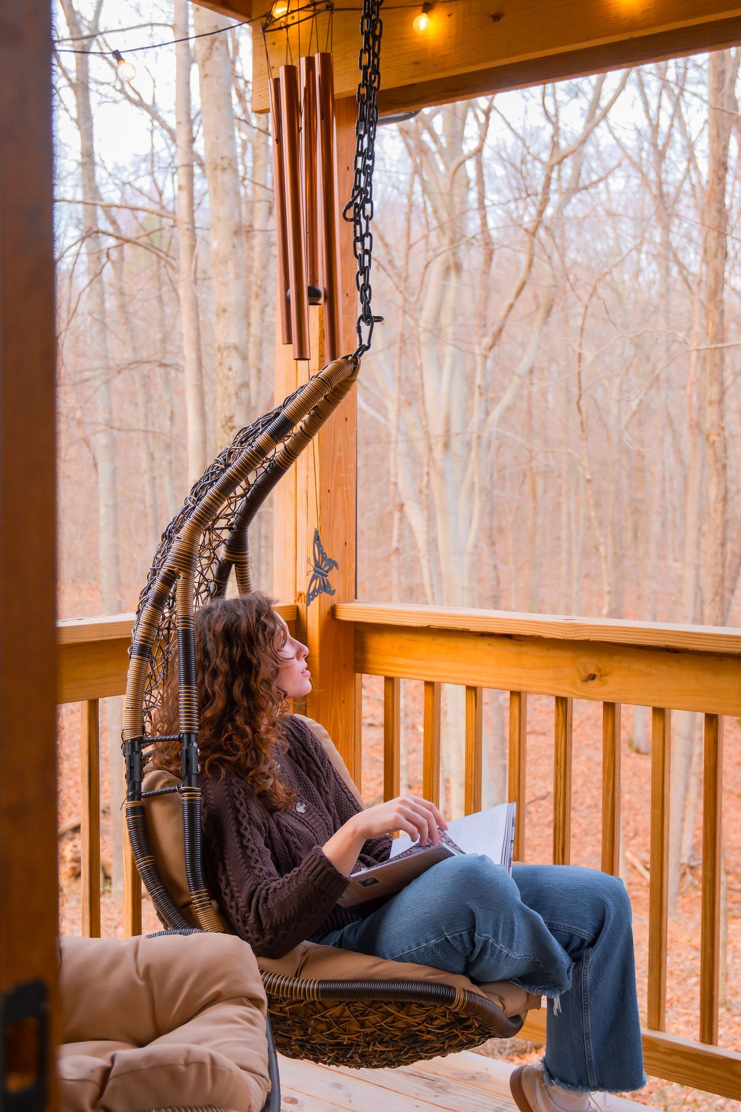 Woman relaxing in hanging wicker chair on wooden porch surrounded by bare winter trees with wind chimes.
